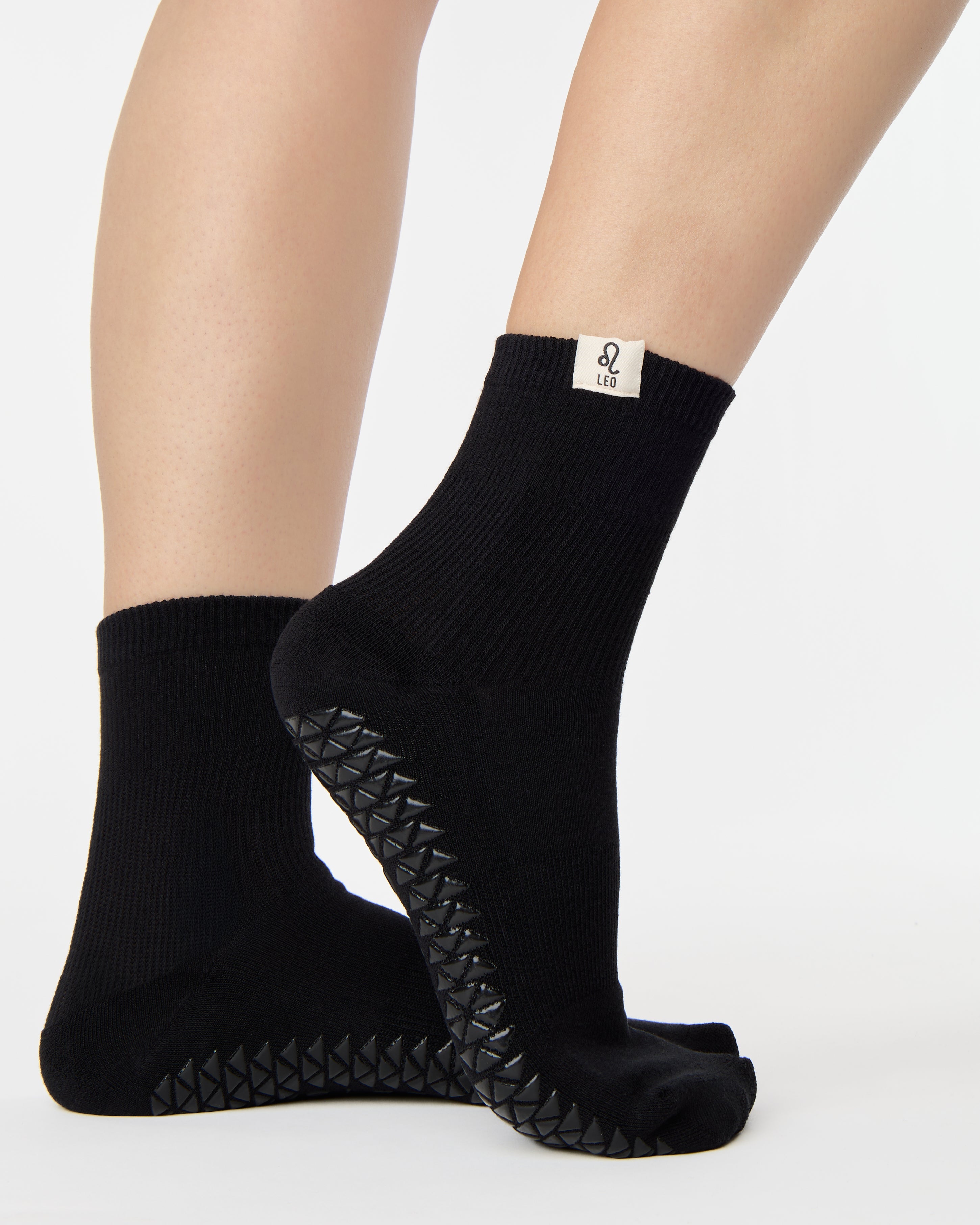 Woman wearing a solid black ankle grip sock with leo zodiac sign embroidered on the side. Made for pilates, barre, yoga, and beyond. 