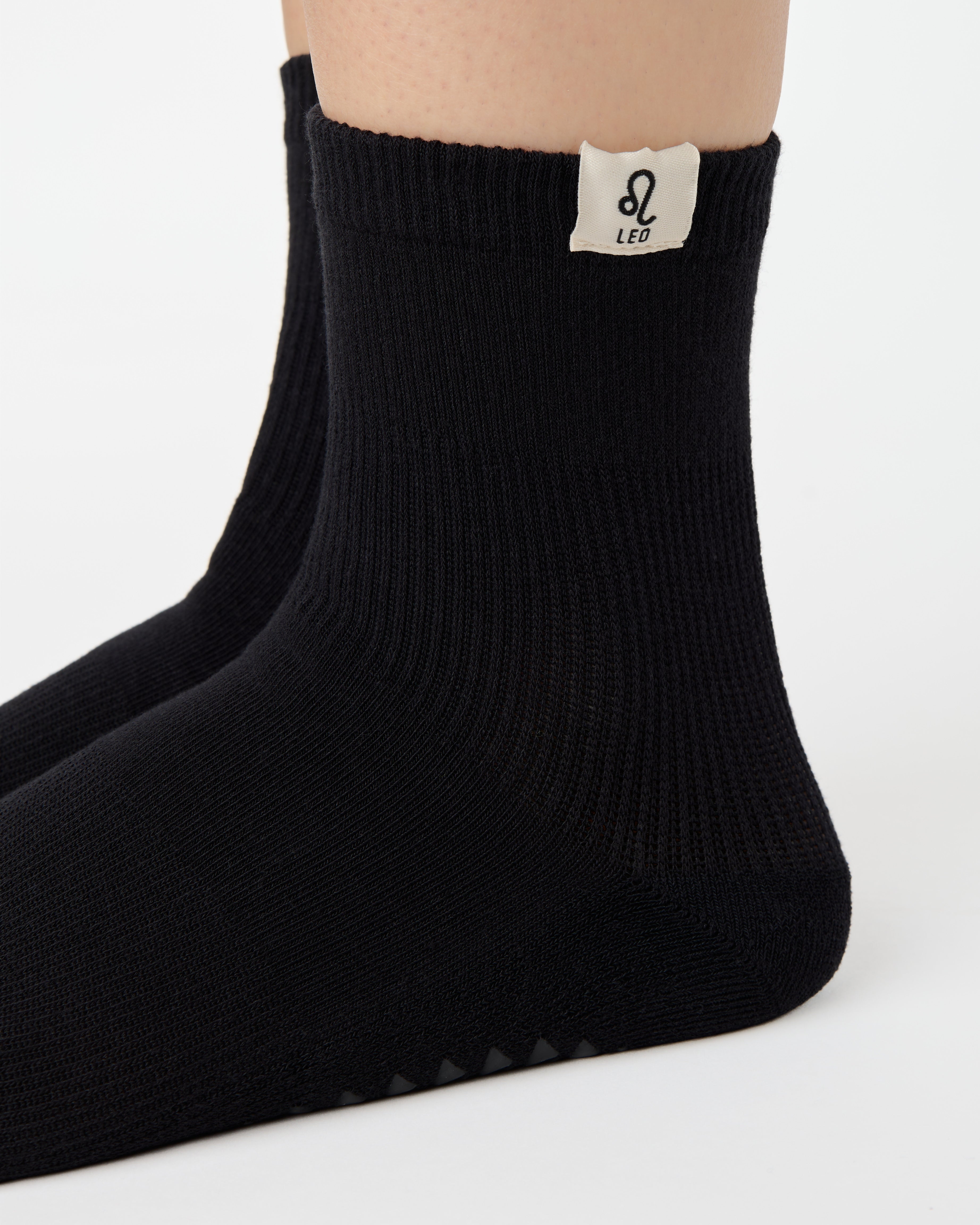 Woman wearing a solid black ankle grip sock with leo zodiac sign embroidered on the side. Made for pilates, barre, yoga, and beyond. 