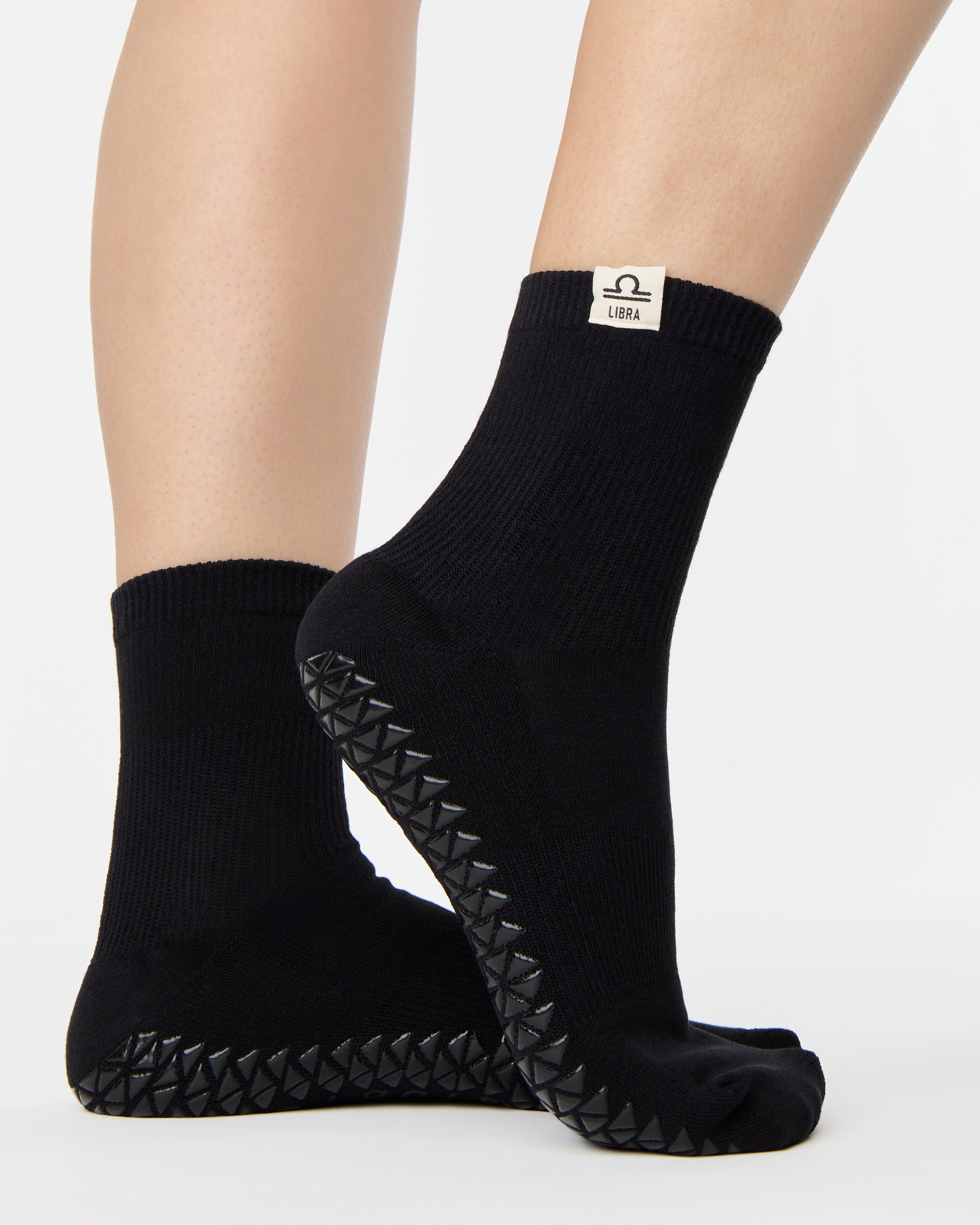 Woman wearing a solid black ankle grip sock with libra zodiac sign embroidered on the side. Made for pilates, barre, yoga, and beyond. 
