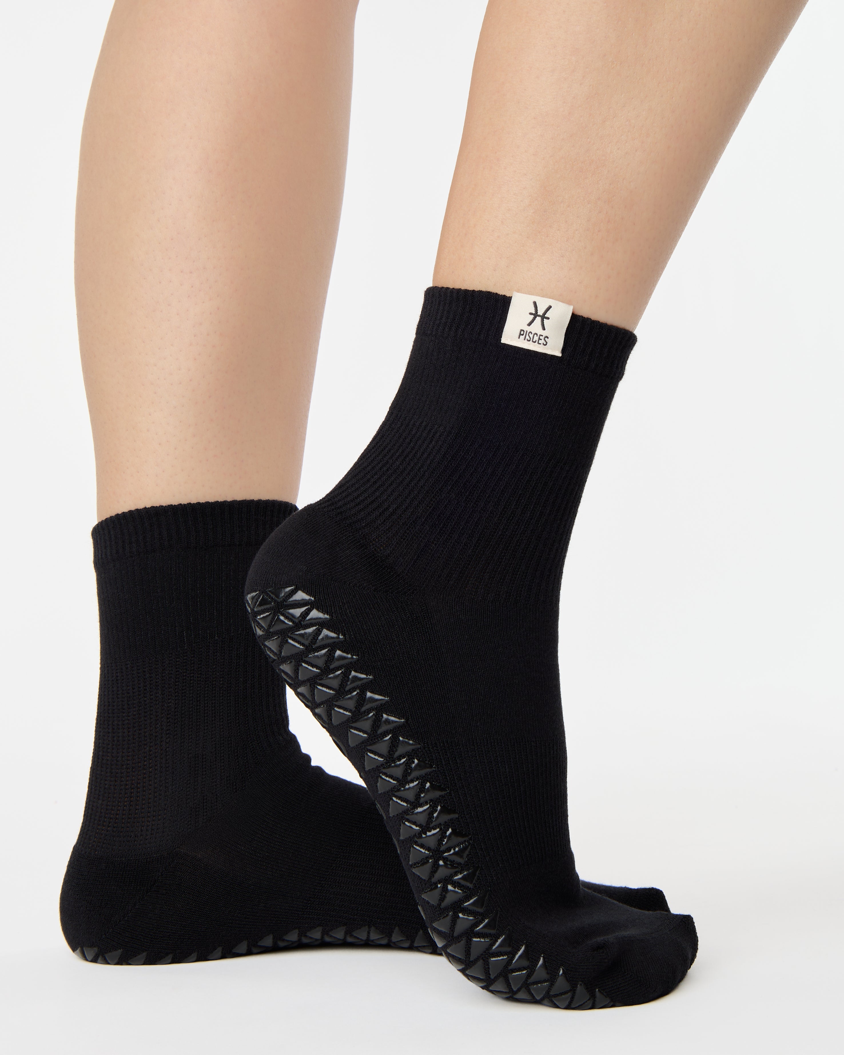 Woman wearing a solid black ankle grip sock with pisces zodiac sign embroidered on the side. Made for pilates, barre, yoga, and beyond. 