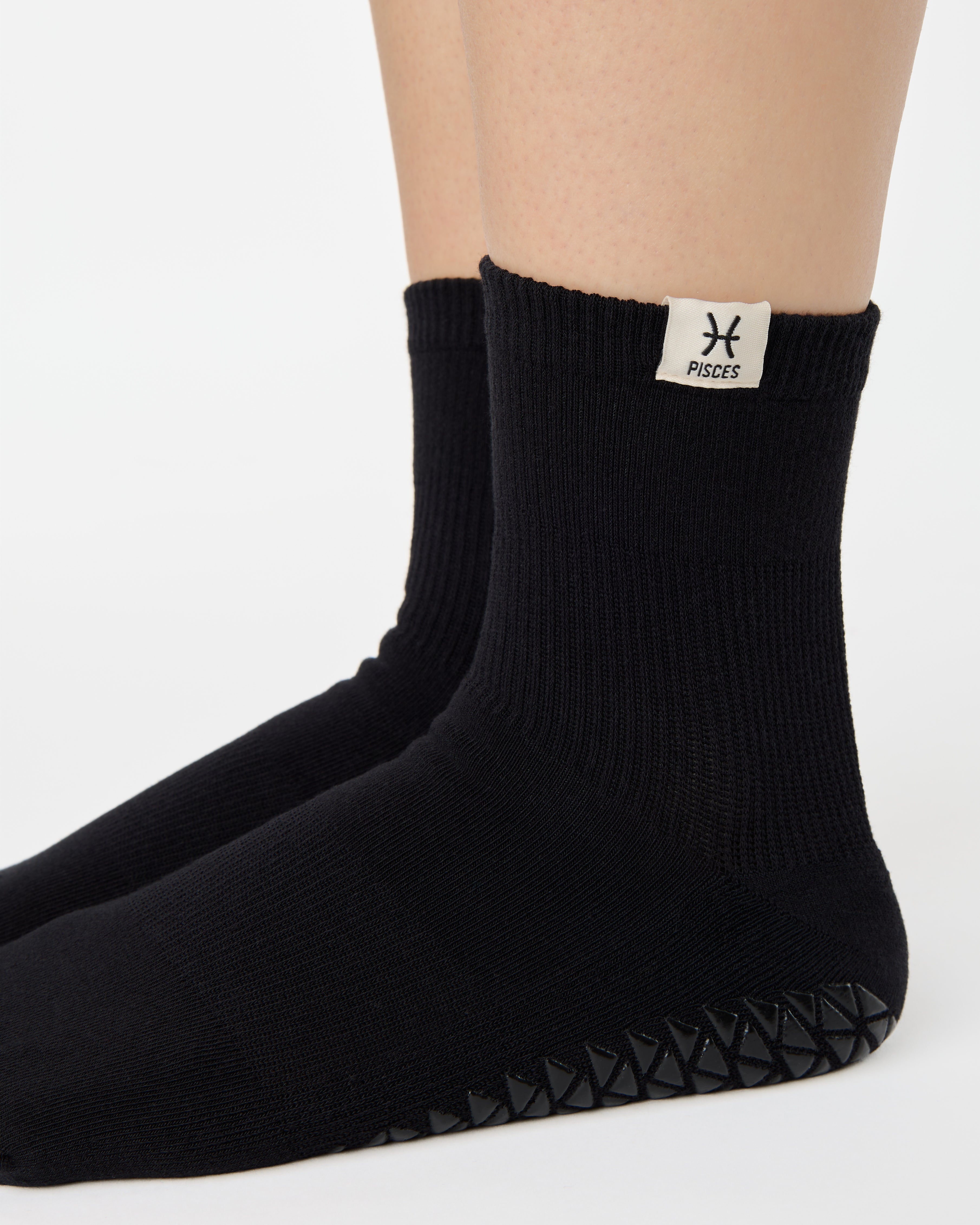 Woman wearing a solid black ankle grip sock with pisces zodiac sign embroidered on the side. Made for pilates, barre, yoga, and beyond. 