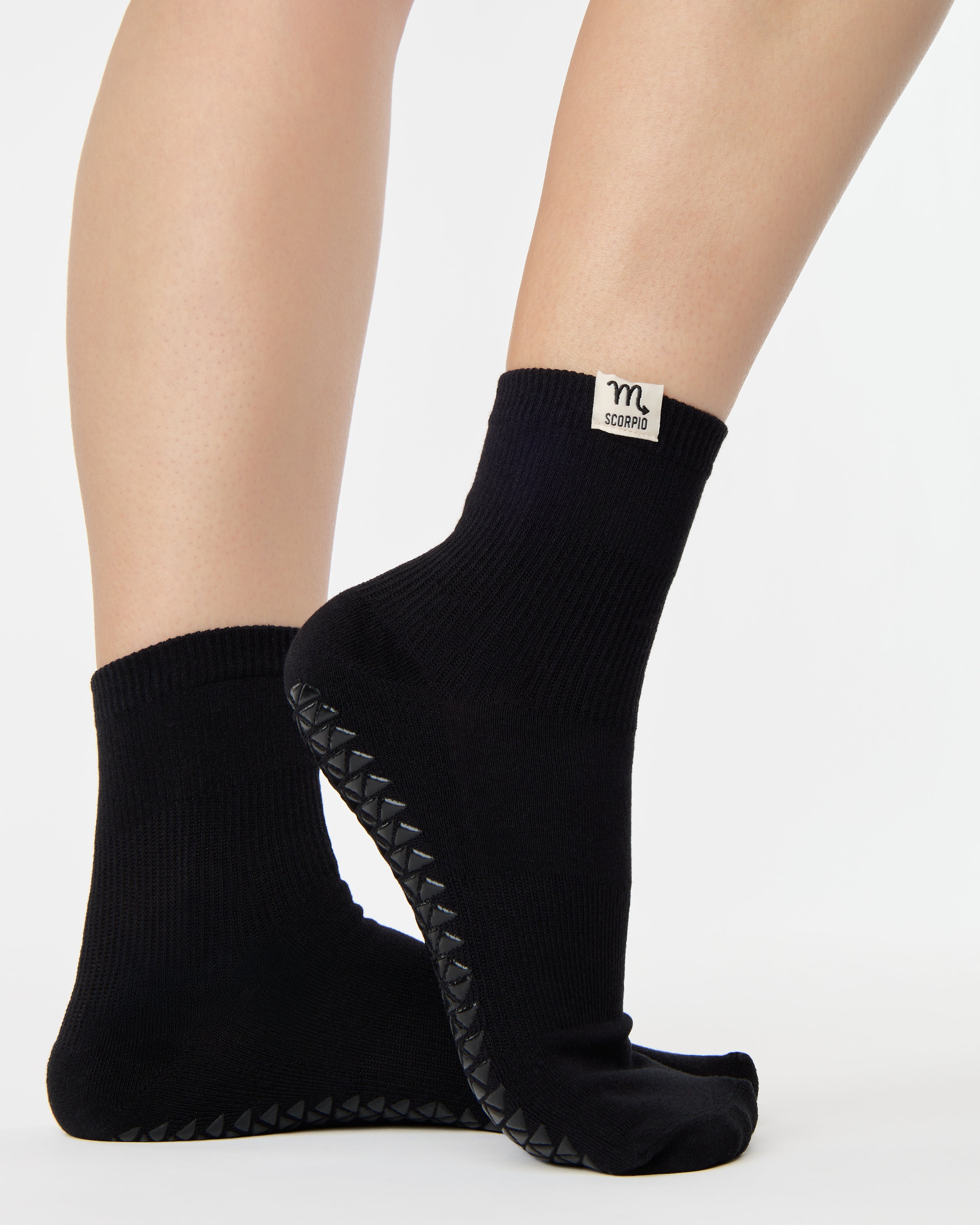 Woman wearing a solid black ankle grip sock with scorpio zodiac sign embroidered on the side. Made for pilates, barre, yoga, and beyond. 