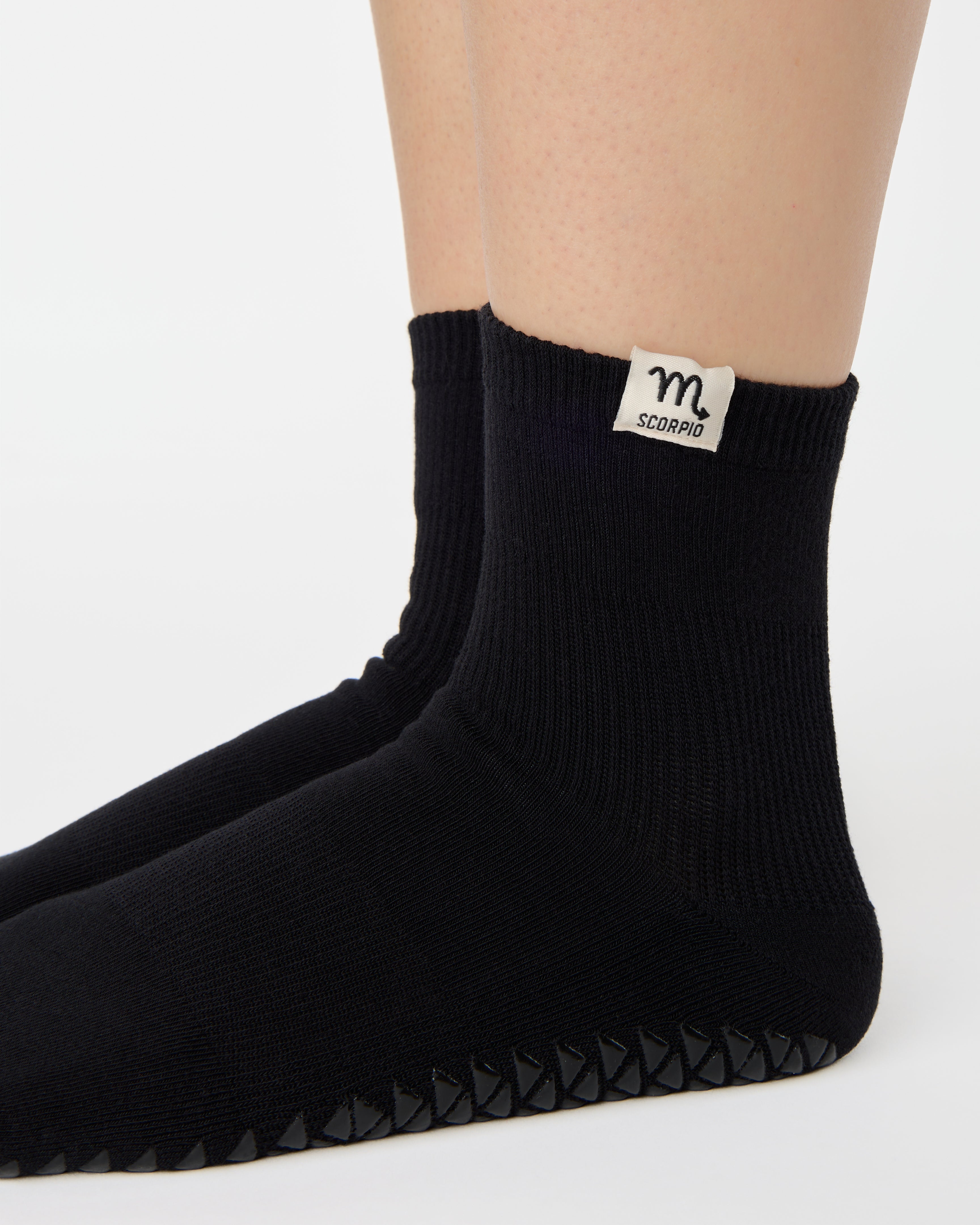 Woman wearing a solid black ankle grip sock with scorpio zodiac sign embroidered on the side. Made for pilates, barre, yoga, and beyond. 