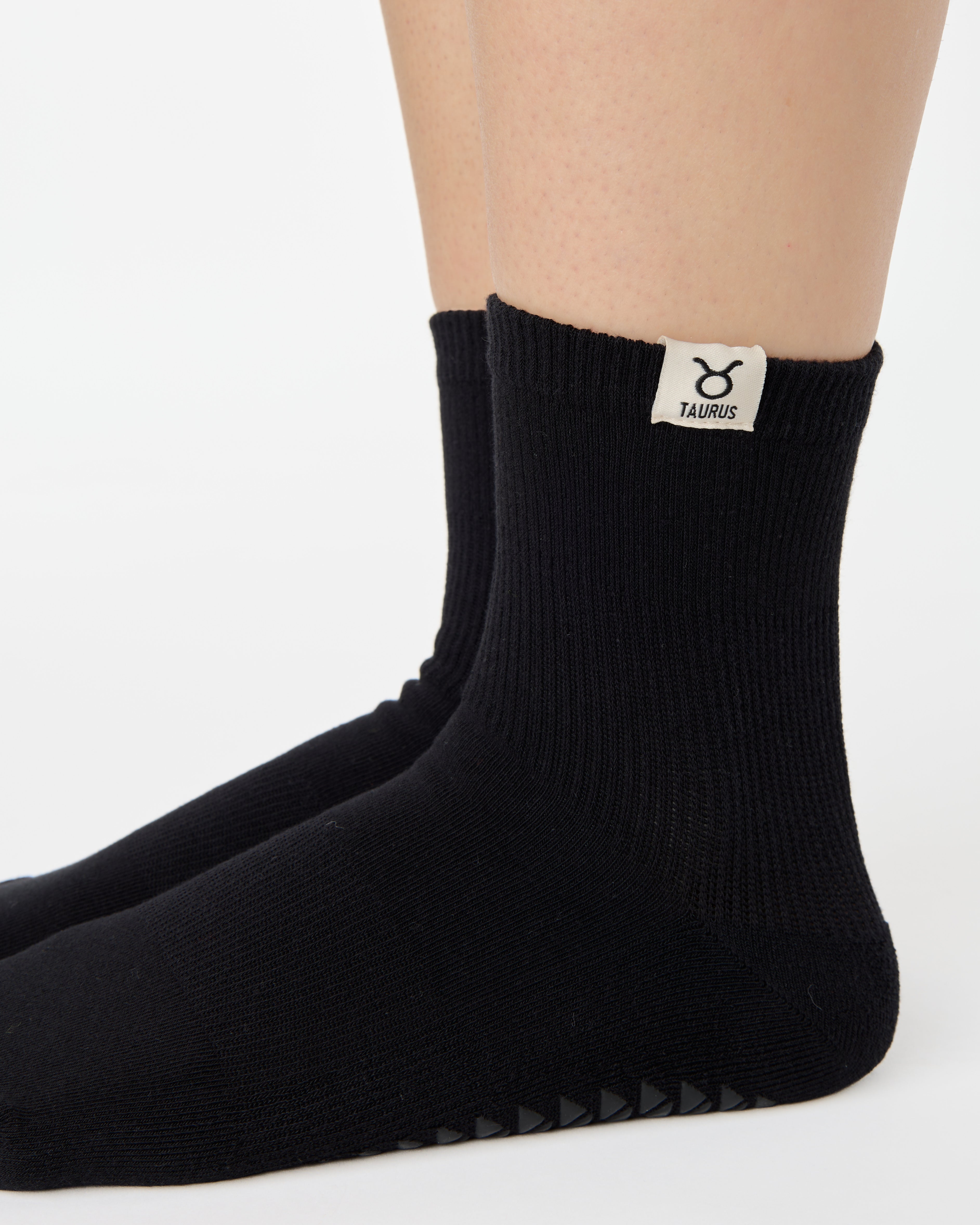 Woman wearing a solid black ankle grip sock with taurus zodiac sign embroidered on the side. Made for pilates, barre, yoga, and beyond. 