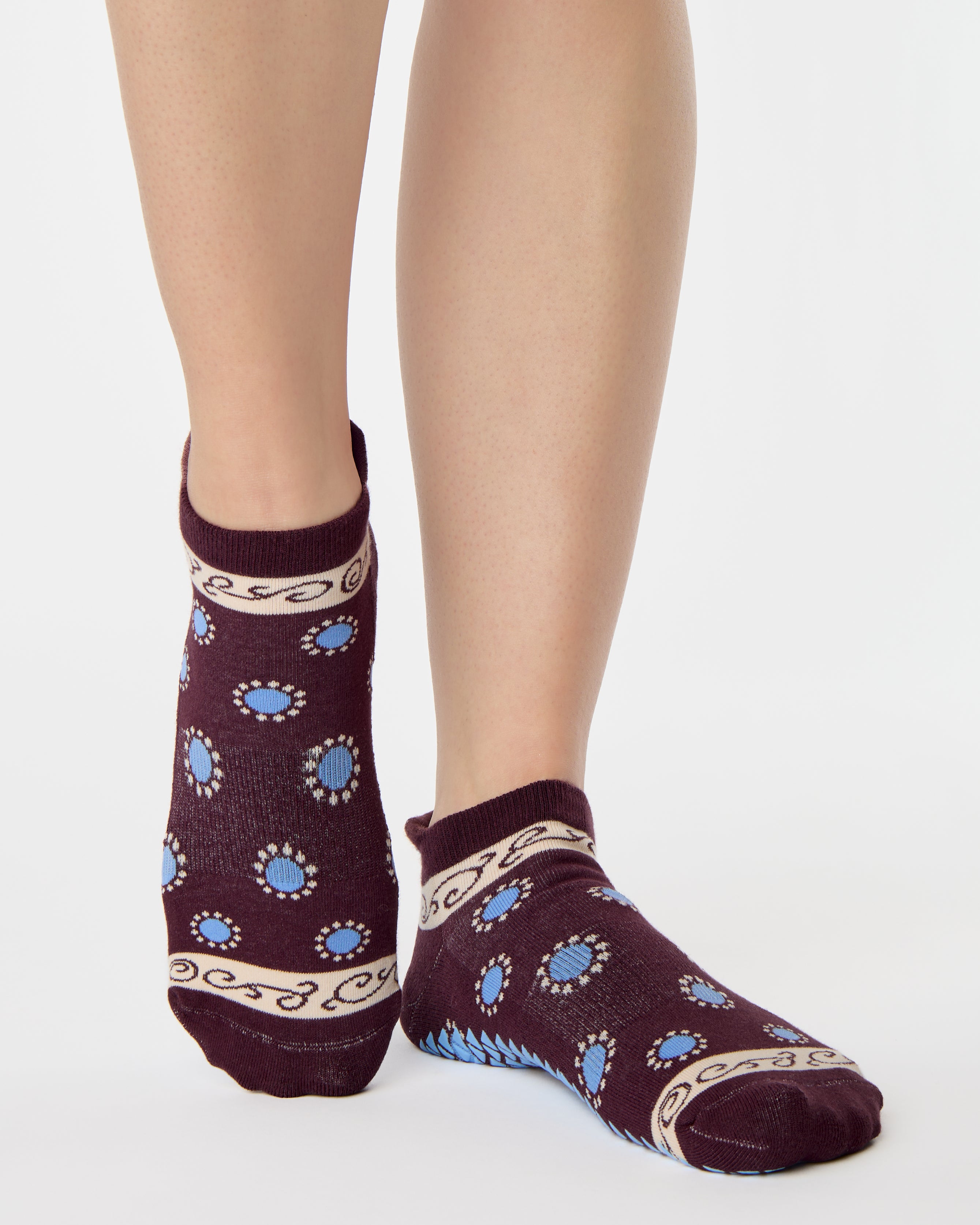 Woman wearing a full foot grip sock that has the design of a classic bandana, in the hues brown cherry and blue and beige detailing. Made for pilates, barre, yoga and beyond. 