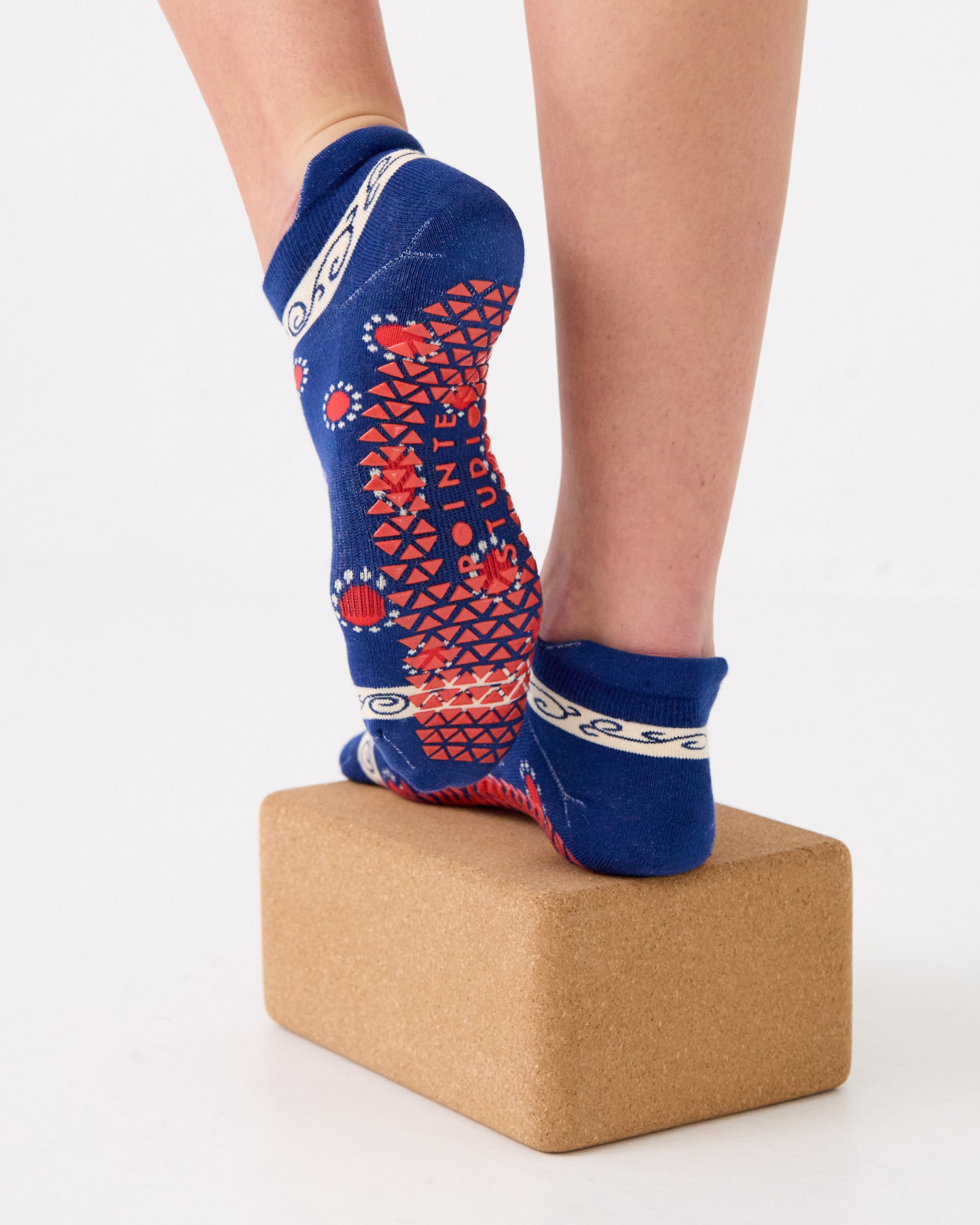 Woman on a yoga block wearing a full foot grip sock that has the design of a classic bandana, in a blue hue with red and white detailing. Made for pilates, barre, yoga and beyond. 