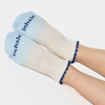 Women wearing an ombre white into light blue full foot grip sock with a navy blue ruffle cuff and inhale and exhale written on the top of the toes, made for pilates, barre, yoga, and beyond.