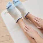 Women on a yoga mat wearing an ombre white into light blue full foot grip sock with a navy blue ruffle cuff and inhale and exhale written on the top of the toes, made for pilates, barre, yoga, and beyond.