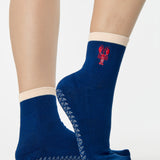 Woman wearing a solid navy blue ankle grip sock with a beige solid cuff and a red lobster embroidered on the ankle. Made for pilates, barre, yoga, and beyond. 