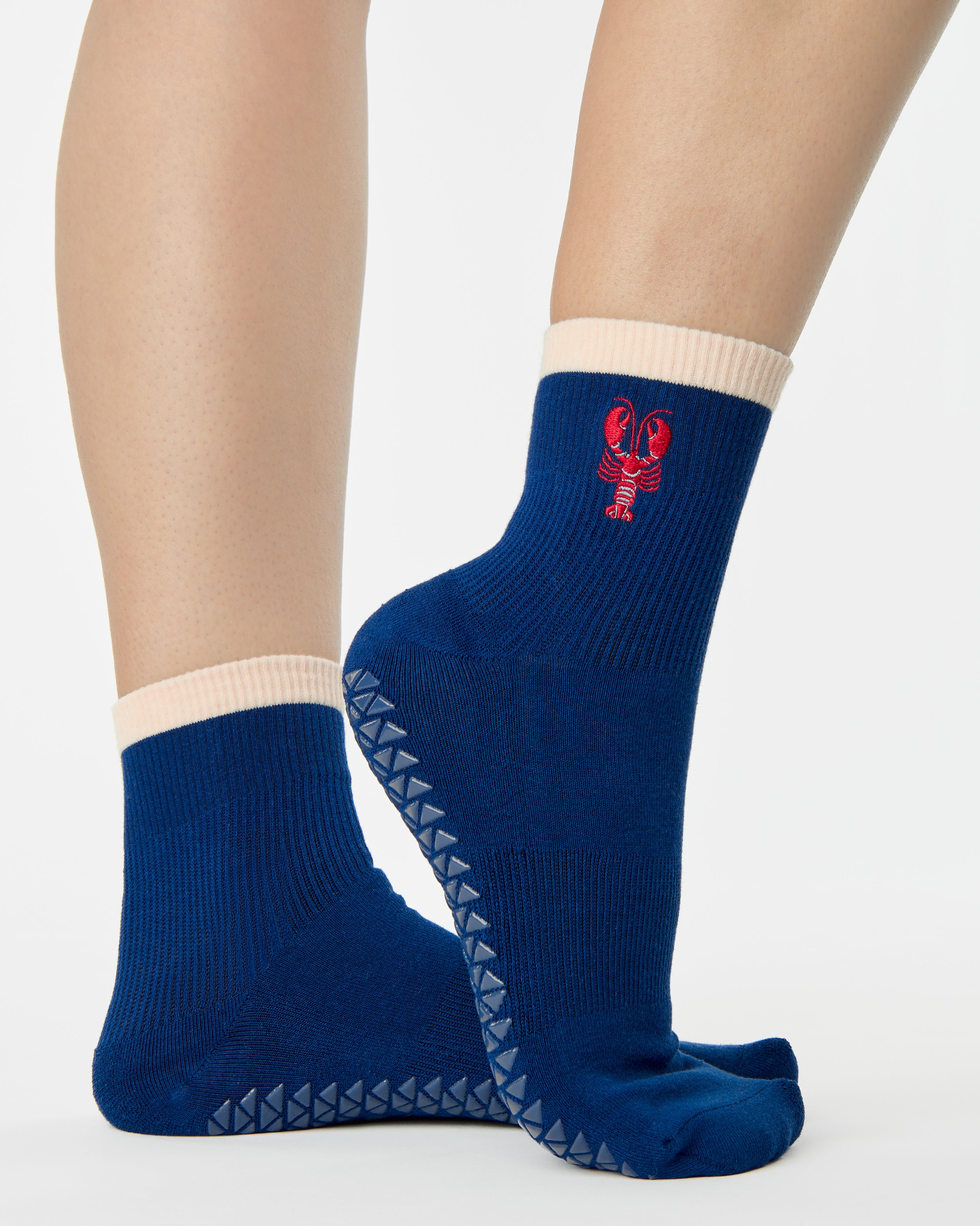 Woman wearing a solid navy blue ankle grip sock with a beige solid cuff and a red lobster embroidered on the ankle. Made for pilates, barre, yoga, and beyond. 