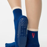 Woman wearing a solid navy blue ankle grip sock with a beige solid cuff and a red lobster embroidered on the ankle. Made for pilates, barre, yoga, and beyond. 