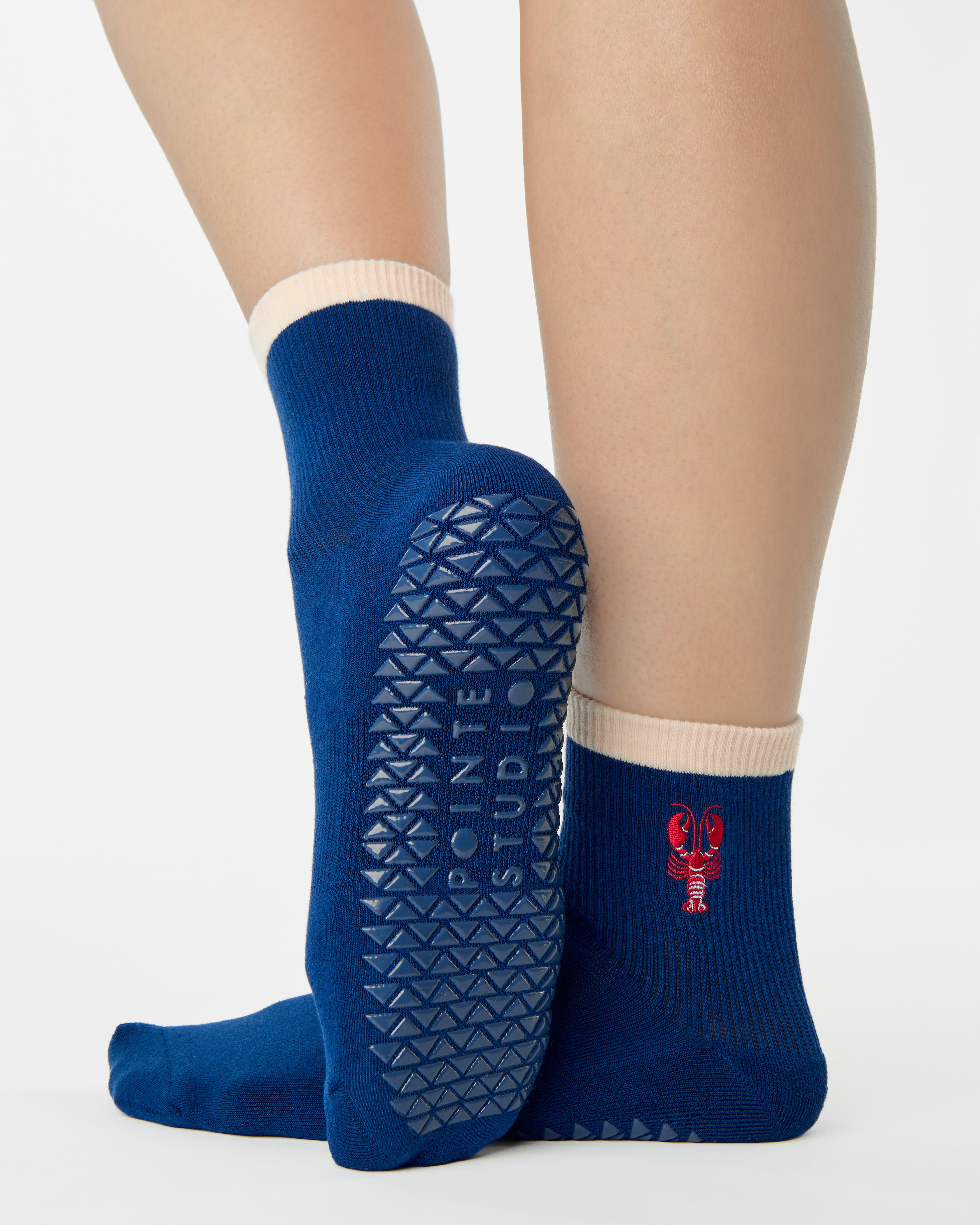 Woman wearing a solid navy blue ankle grip sock with a beige solid cuff and a red lobster embroidered on the ankle. Made for pilates, barre, yoga, and beyond. 