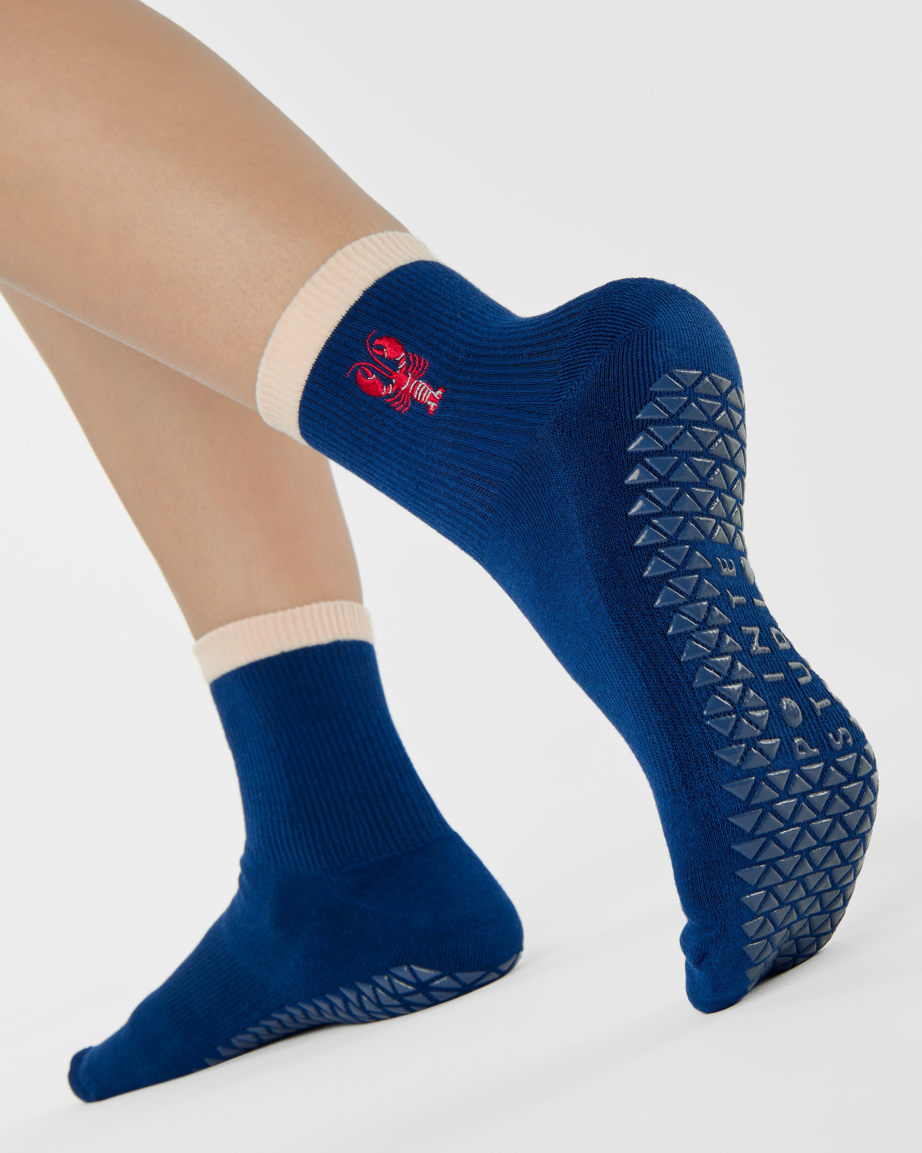 Woman wearing a solid navy blue ankle grip sock with a beige solid cuff and a red lobster embroidered on the ankle. Made for pilates, barre, yoga, and beyond. 