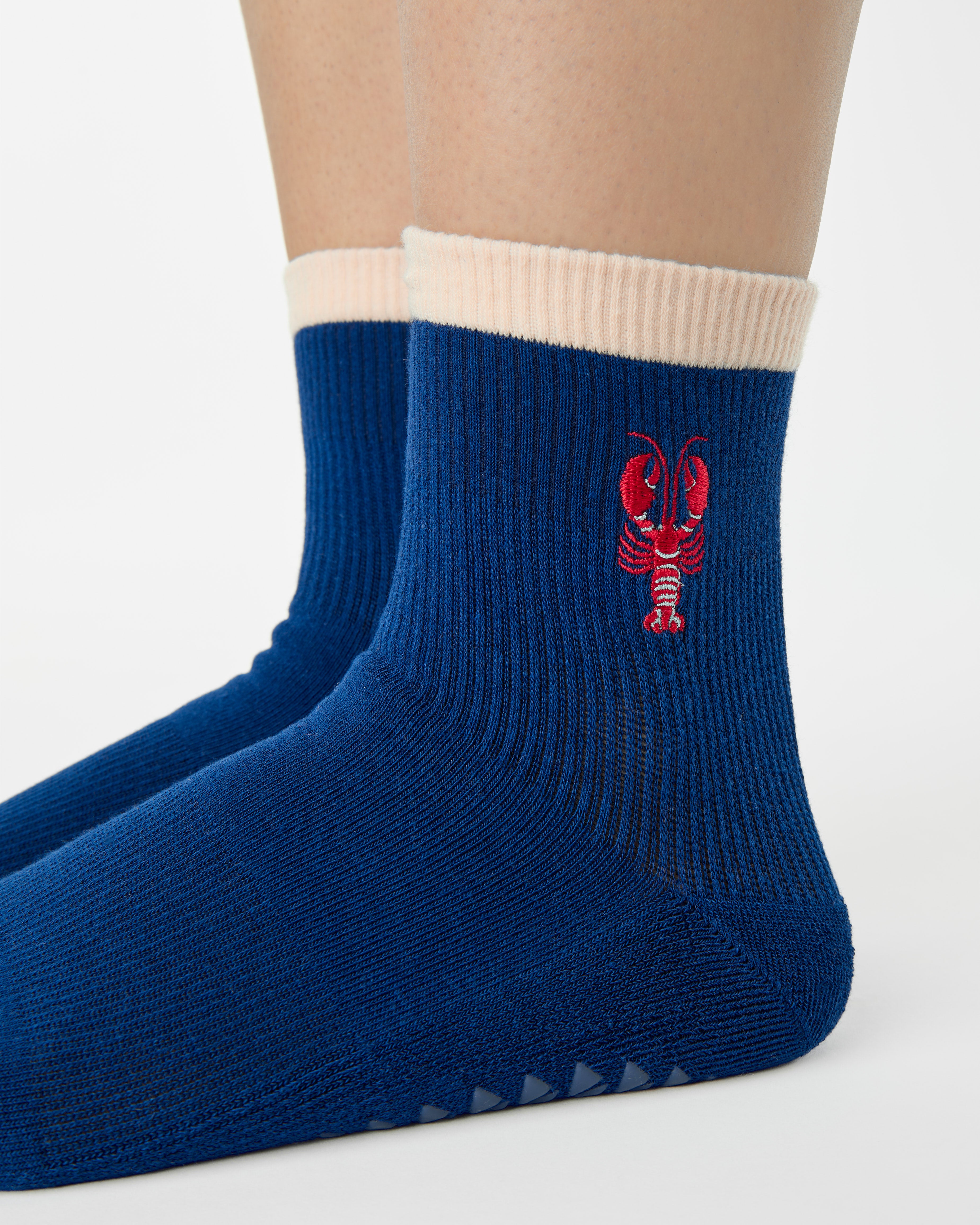 Woman wearing a solid navy blue ankle grip sock with a beige solid cuff and a red lobster embroidered on the ankle. Made for pilates, barre, yoga, and beyond. 