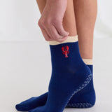 Woman wearing a solid navy blue ankle grip sock with a beige solid cuff and a red lobster embroidered on the ankle. Made for pilates, barre, yoga, and beyond. 