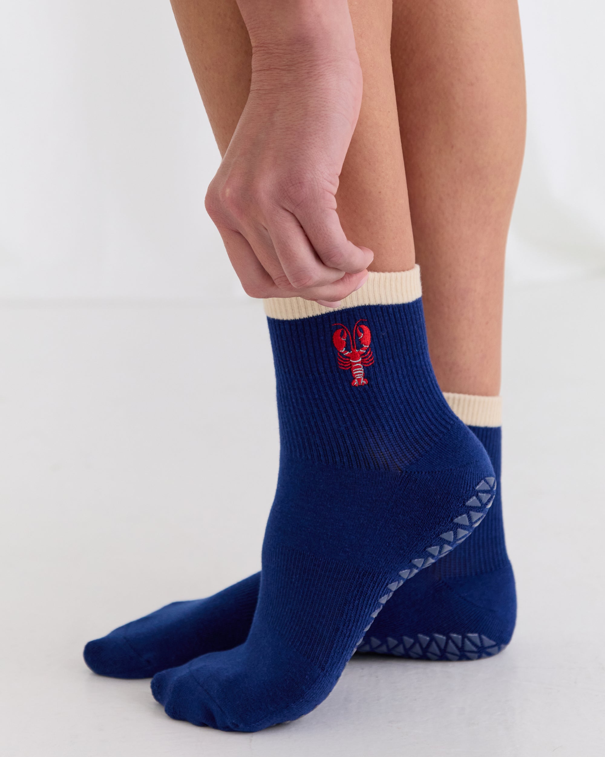 Woman wearing a solid navy blue ankle grip sock with a beige solid cuff and a red lobster embroidered on the ankle. Made for pilates, barre, yoga, and beyond. 