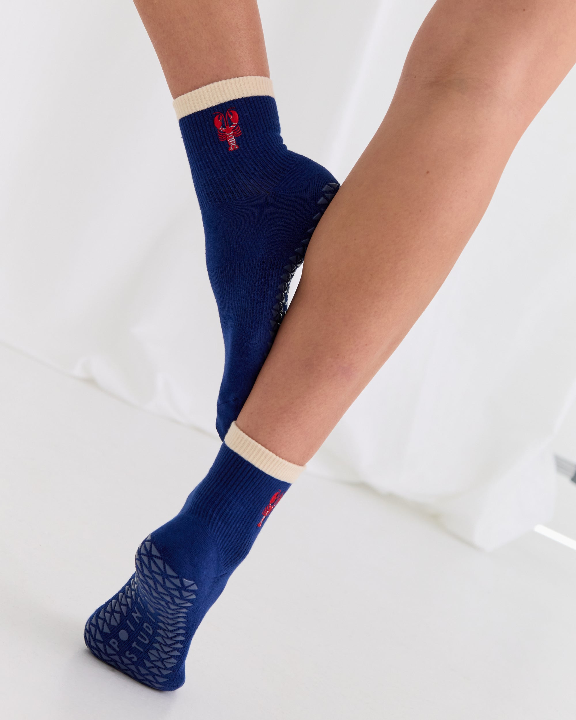 Woman wearing a solid navy blue ankle grip sock with a beige solid cuff and a red lobster embroidered on the ankle. Made for pilates, barre, yoga, and beyond. 