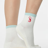 Woman wearing a solid white ankle grip sock with a light blue solid cuff and a red shrimp embroidered on the ankle. Made for pilates, barre, yoga, and beyond. 