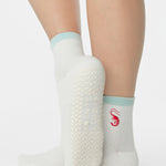 Woman wearing a solid white ankle grip sock with a light blue solid cuff and a red shrimp embroidered on the ankle. Made for pilates, barre, yoga, and beyond. 