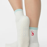 Woman wearing a solid white ankle grip sock with a light blue solid cuff and a red shrimp embroidered on the ankle. Made for pilates, barre, yoga, and beyond. 