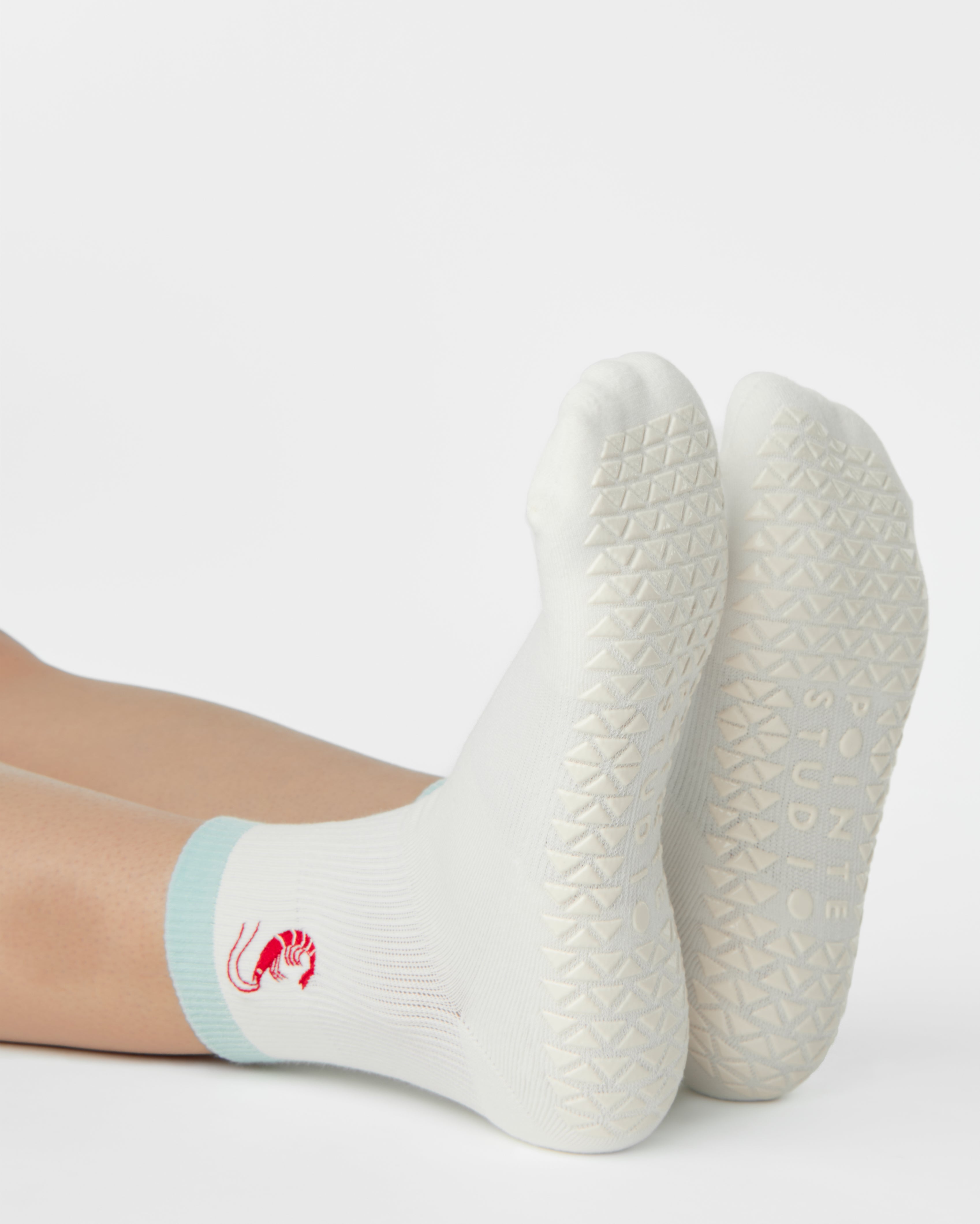 Woman wearing a solid white ankle grip sock with a light blue solid cuff and a red shrimp embroidered on the ankle. Made for pilates, barre, yoga, and beyond. 