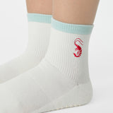 Woman wearing a solid white ankle grip sock with a light blue solid cuff and a red shrimp embroidered on the ankle. Made for pilates, barre, yoga, and beyond. 