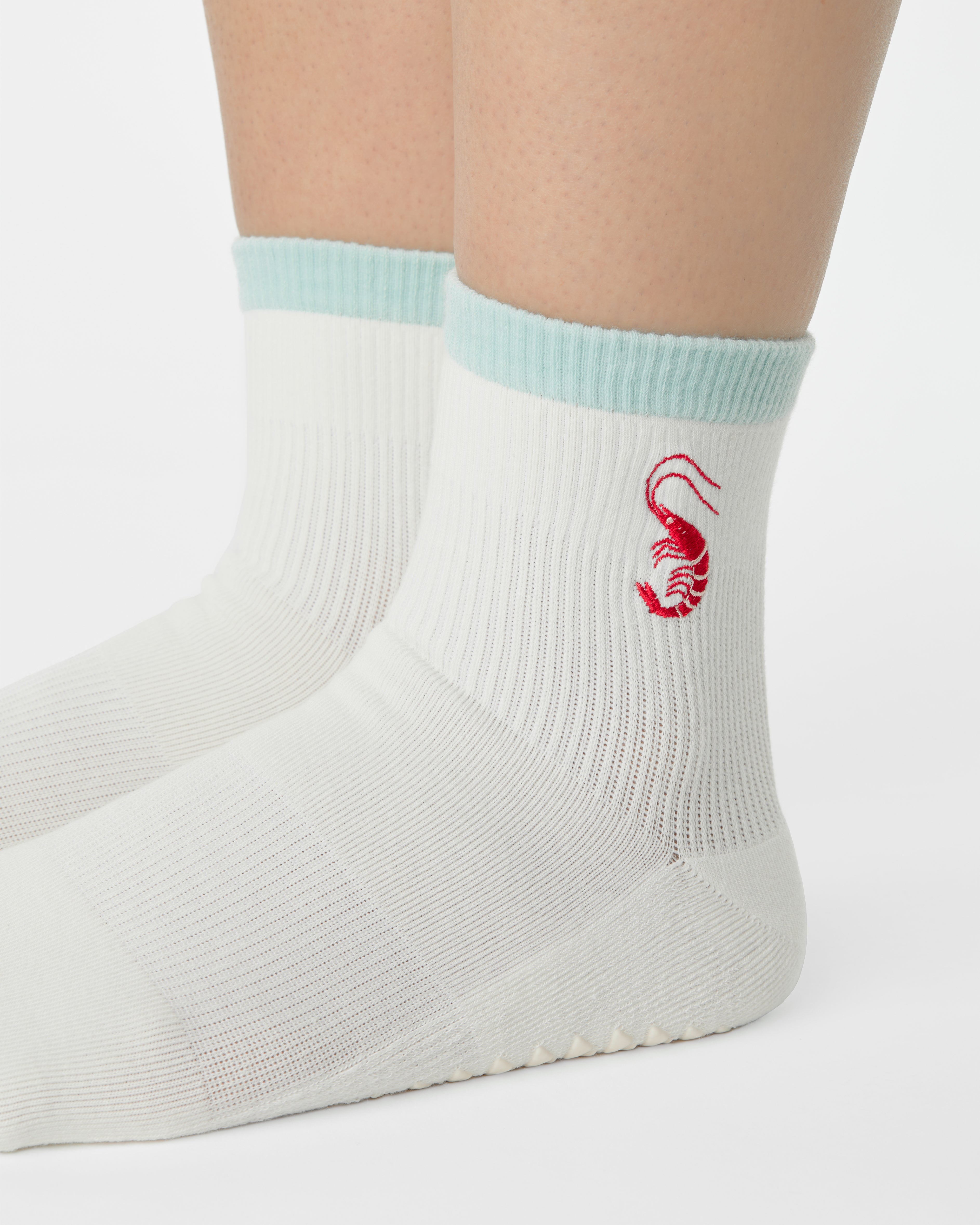 Woman wearing a solid white ankle grip sock with a light blue solid cuff and a red shrimp embroidered on the ankle. Made for pilates, barre, yoga, and beyond. 
