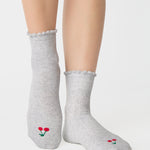 Women wearing a solid heather grey ankle grip sock with a ruffle cuff and with a red cherry on the top of the toes, made for pilates, barre, yoga, and beyond.