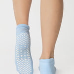 Women wearing a solid white full foot grip sock with a blue evil eye motiff on the top of the toes, made for pilates, barre, yoga, and beyond.