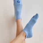 Women wearing a solid white full foot grip sock with a blue evil eye motiff on the top of the toes, made for pilates, barre, yoga, and beyond.