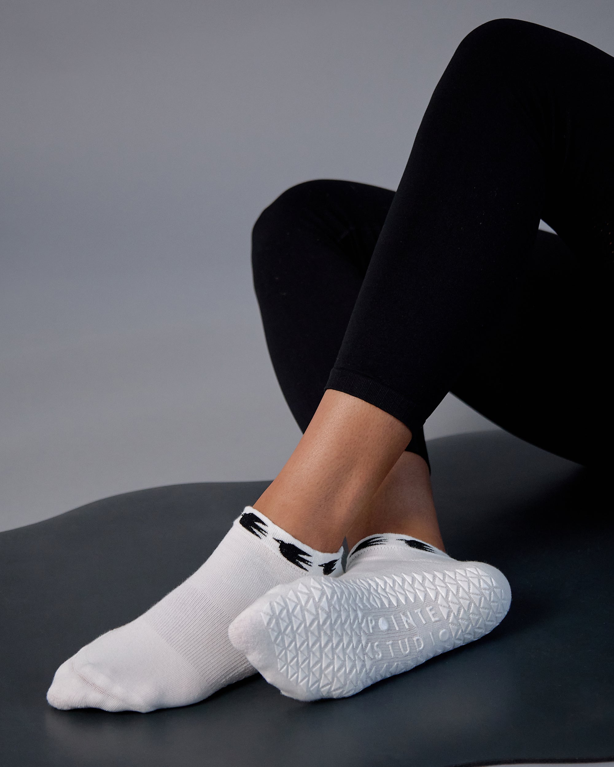 Woman on yoga mat wearing a white sold color full foot grip sock with crows circling the cuff. Made for pilates, barre, yoga, and beyond.