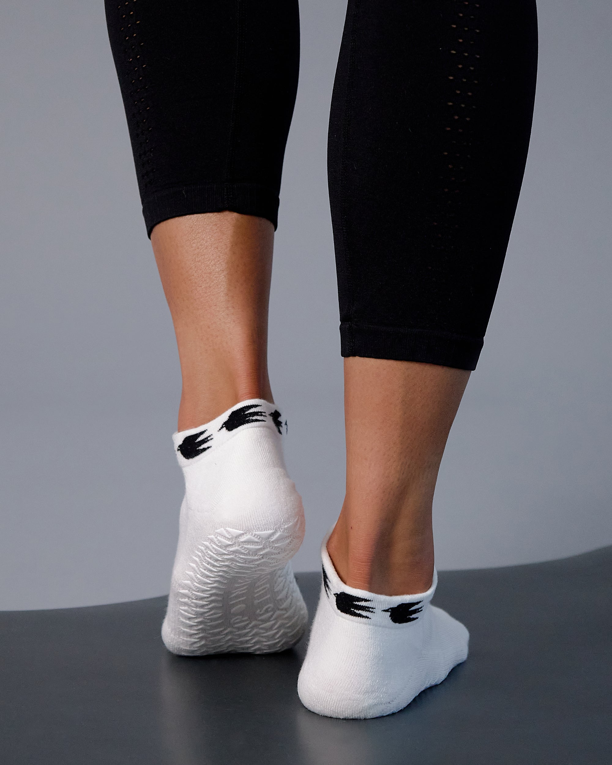 Woman on yoga mat wearing a white sold color full foot grip sock with crows circling the cuff. Made for pilates, barre, yoga, and beyond.