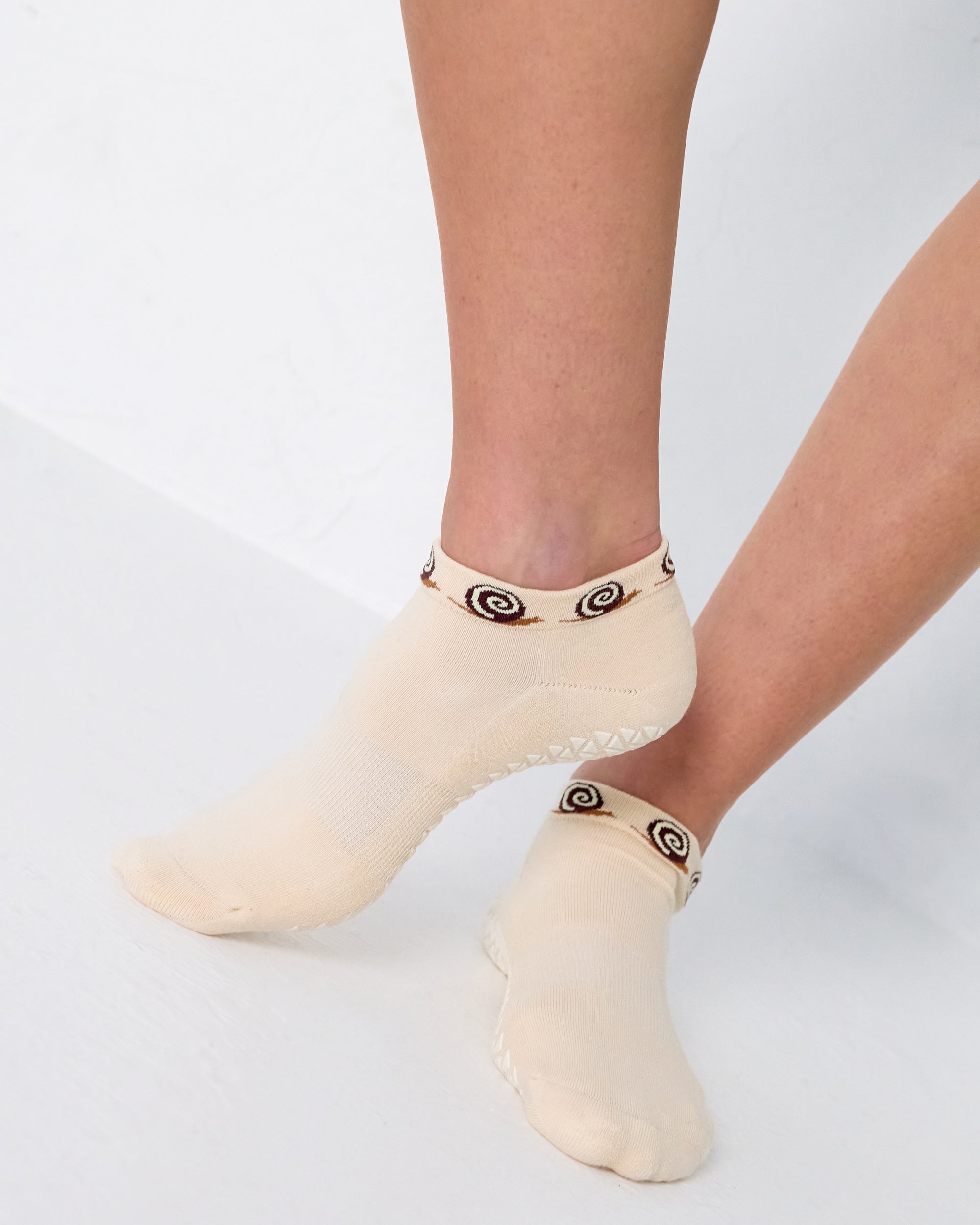 Woman wearing a beige sold color full foot grip sock with snails circling the cuff. Made for pilates, barre, yoga, and beyond.