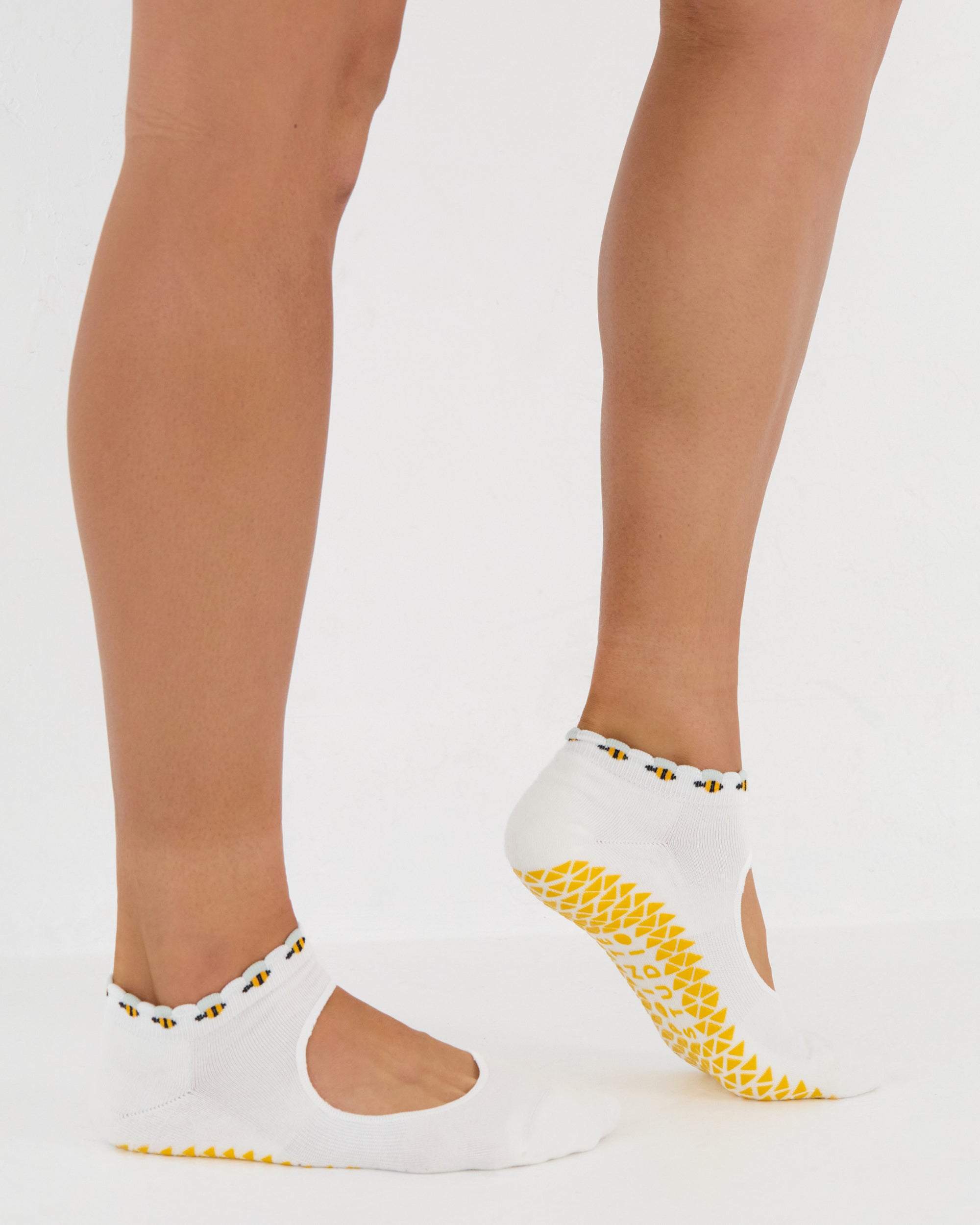 Woman wearing solid white strap grip sock with bumble bees wrapping around the scalloped cuff, made for pilates, barre, yoga, and beyond. 