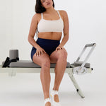 Woman on a pilates reformer wearing solid white strap grip sock with bumble bees wrapping around the scalloped cuff, made for pilates, barre, yoga, and beyond. 