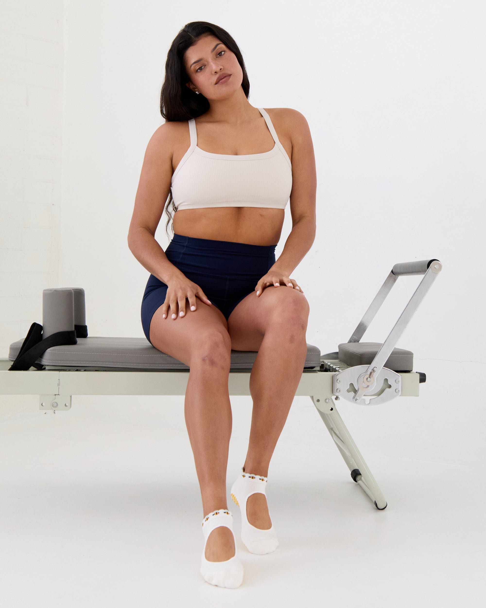 Woman on a pilates reformer wearing solid white strap grip sock with bumble bees wrapping around the scalloped cuff, made for pilates, barre, yoga, and beyond. 