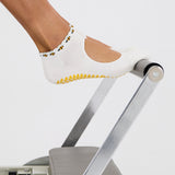 Woman on a pilates reformer wearing solid white strap grip sock with bumble bees wrapping around the scalloped cuff, made for pilates, barre, yoga, and beyond. 