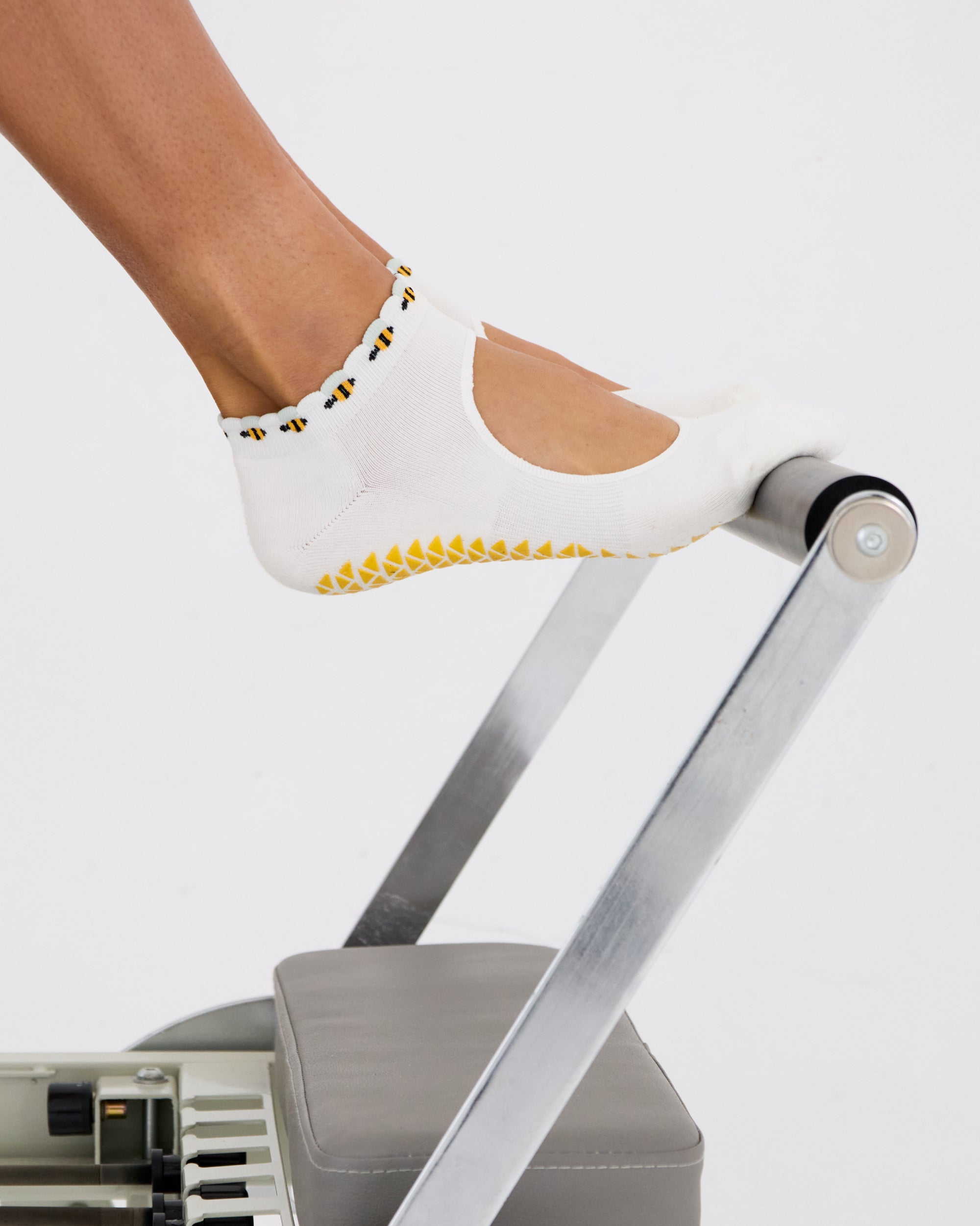 Woman on a pilates reformer wearing solid white strap grip sock with bumble bees wrapping around the scalloped cuff, made for pilates, barre, yoga, and beyond. 