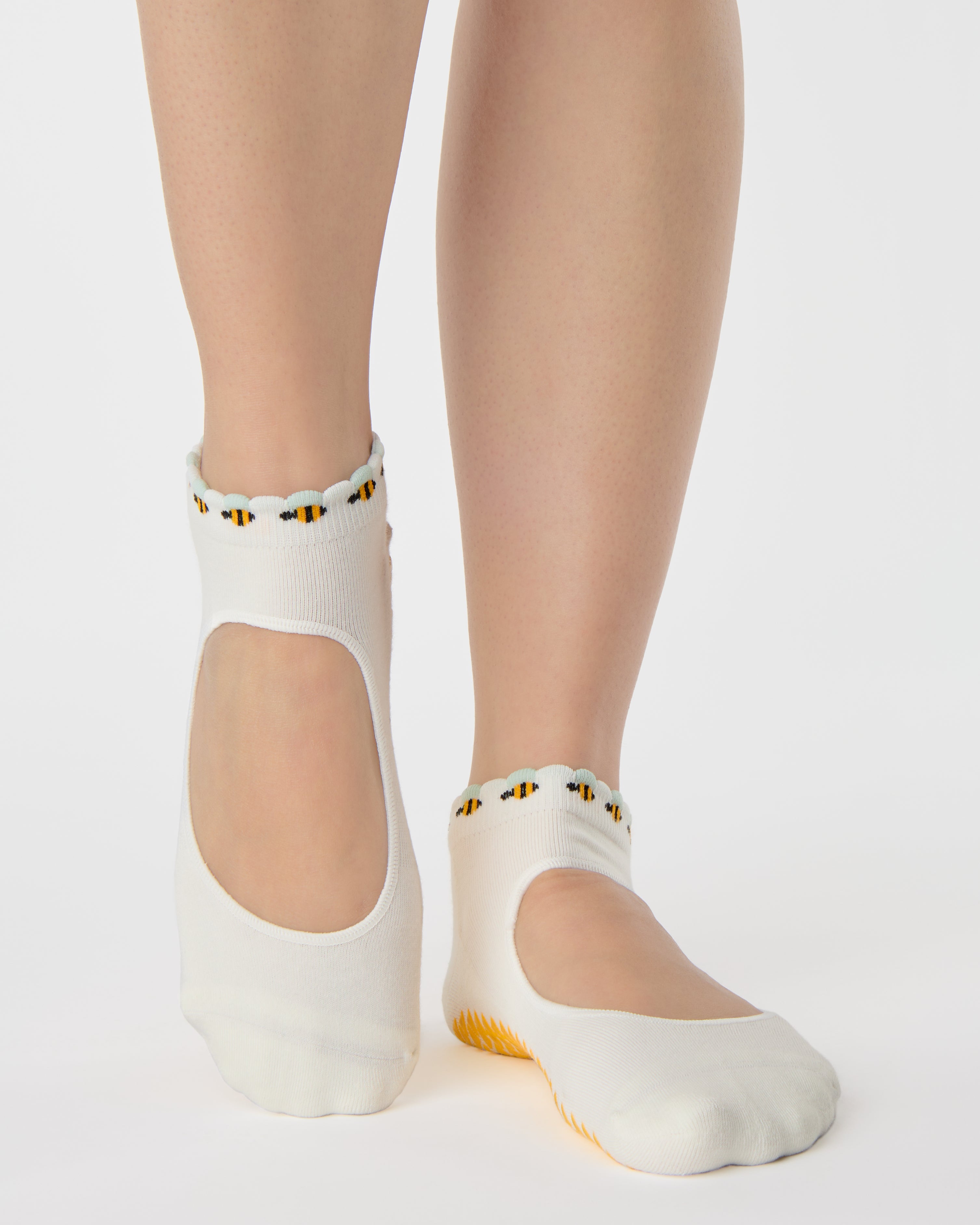 Woman wearing solid white strap grip sock with bumble bees wrapping around the scalloped cuff, made for pilates, barre, yoga, and beyond. 