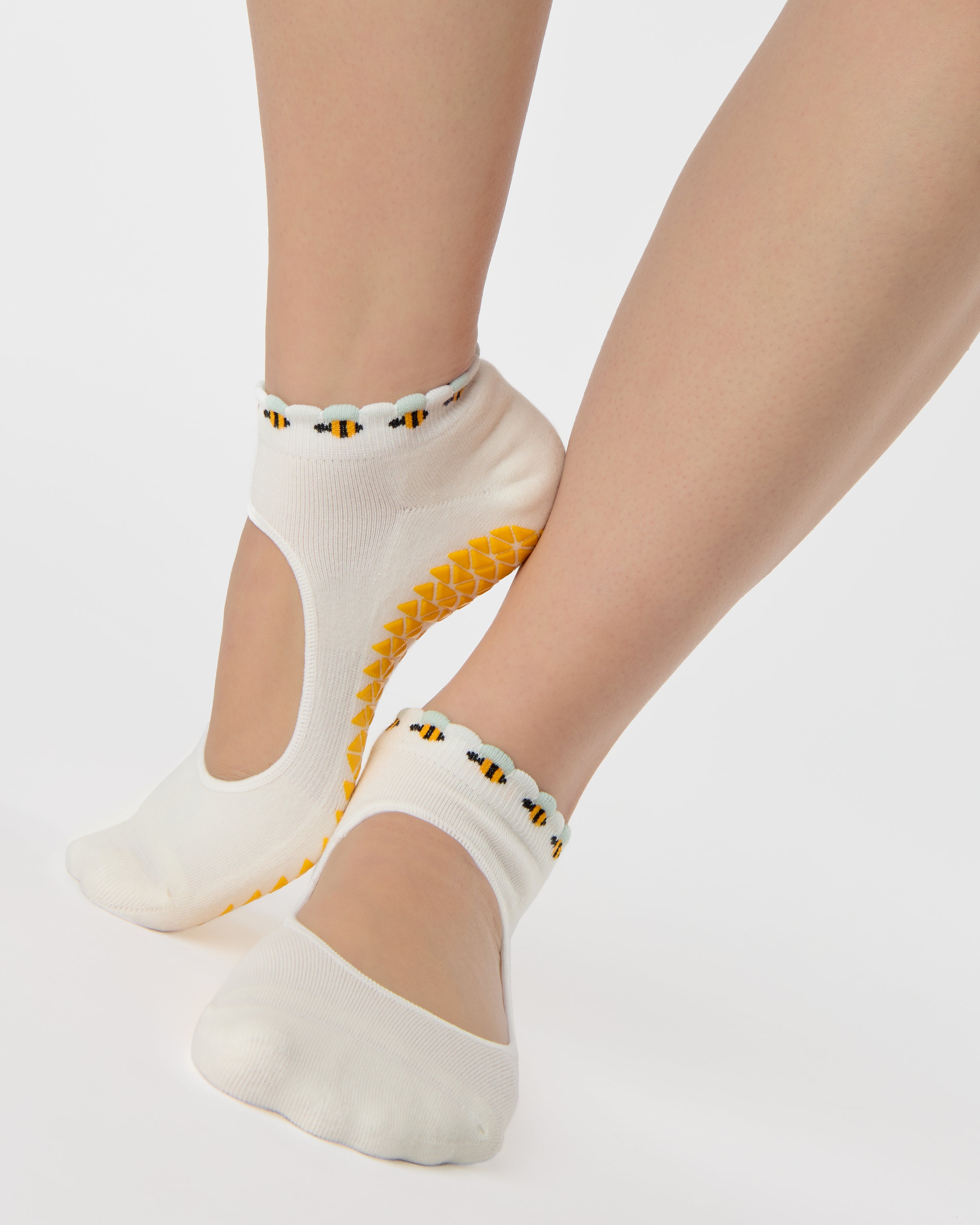 Woman wearing solid white strap grip sock with bumble bees wrapping around the scalloped cuff, made for pilates, barre, yoga, and beyond. 