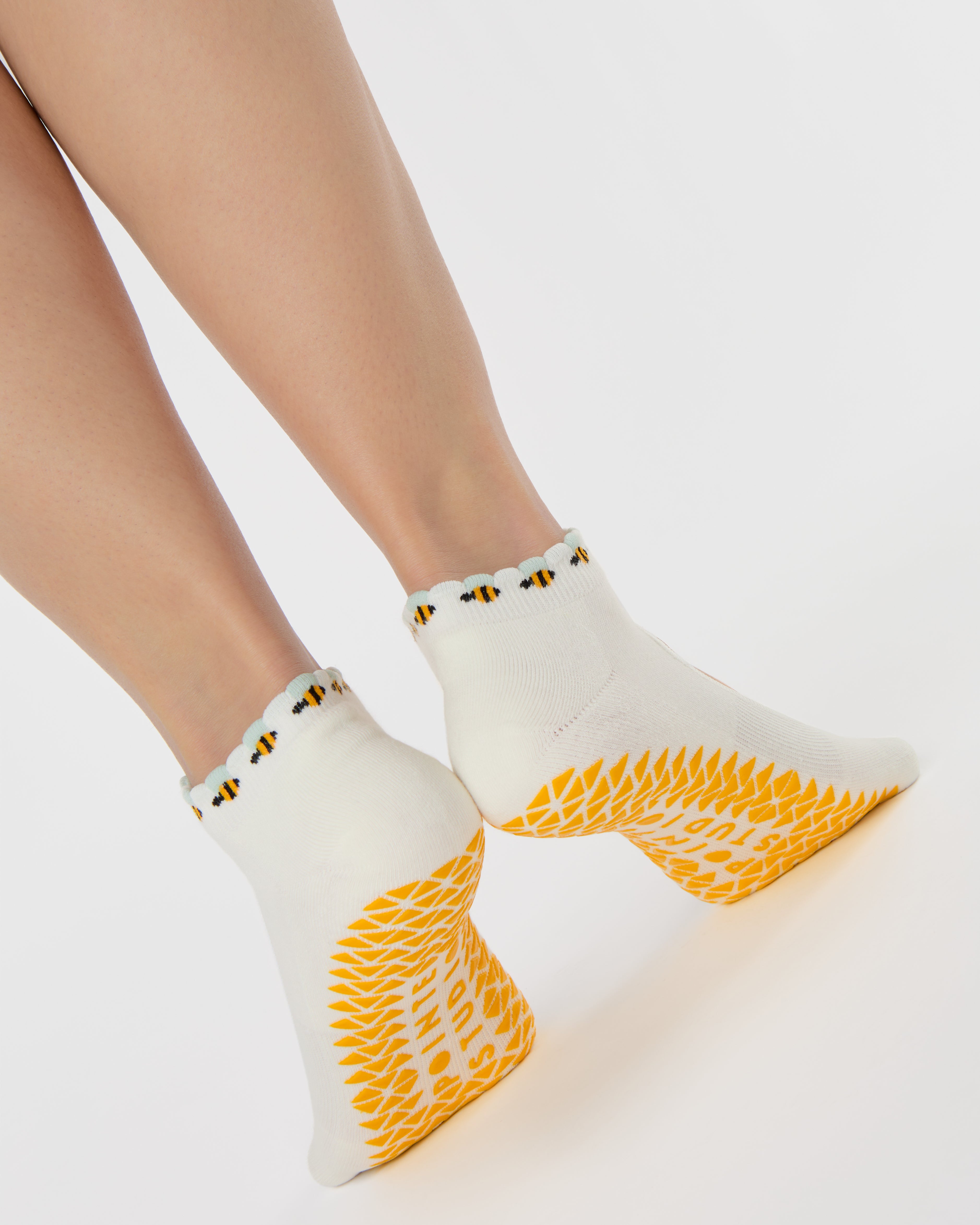 Woman wearing solid white strap grip sock with bumble bees wrapping around the scalloped cuff, made for pilates, barre, yoga, and beyond. 