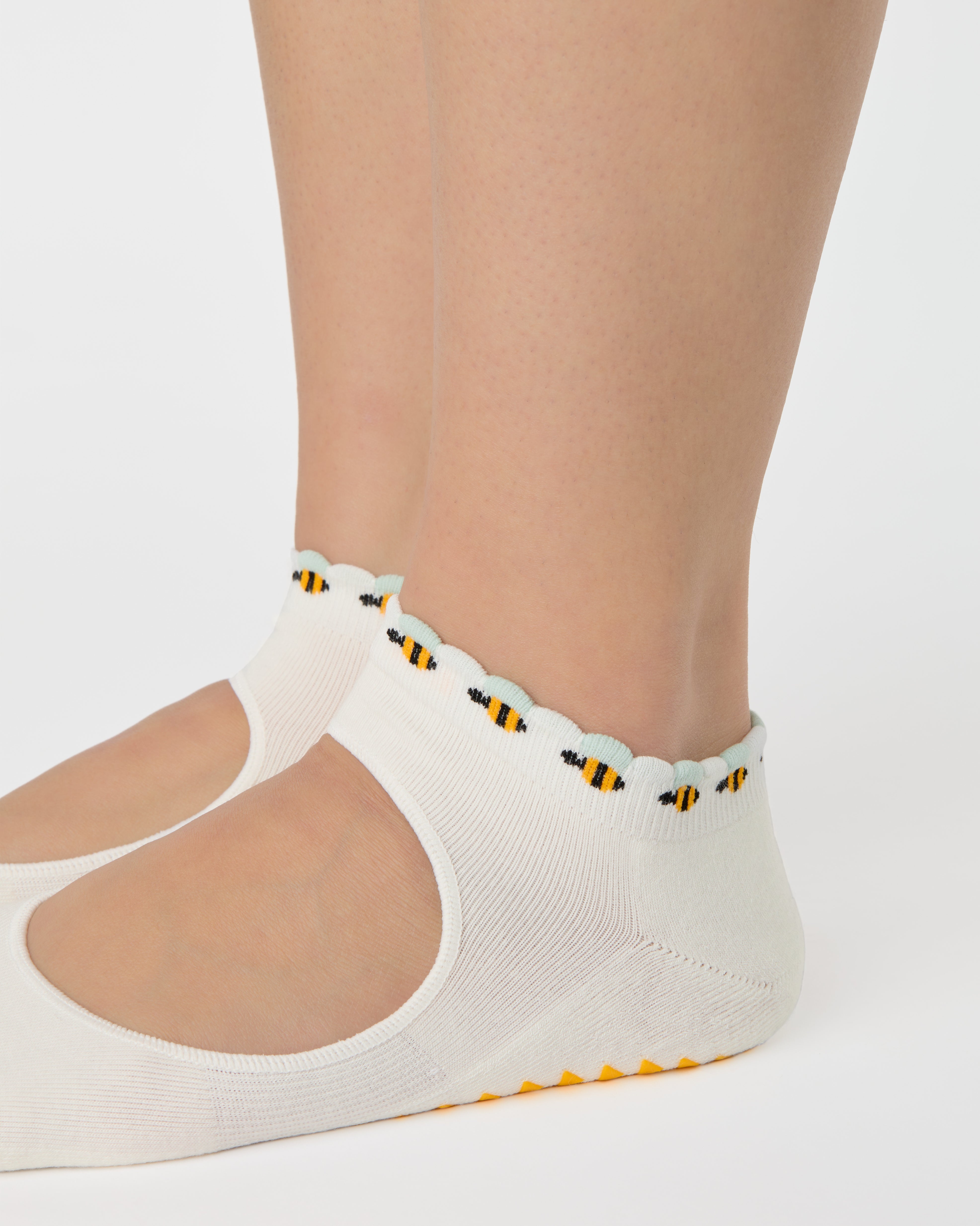 Woman wearing solid white strap grip sock with bumble bees wrapping around the scalloped cuff, made for pilates, barre, yoga, and beyond. 