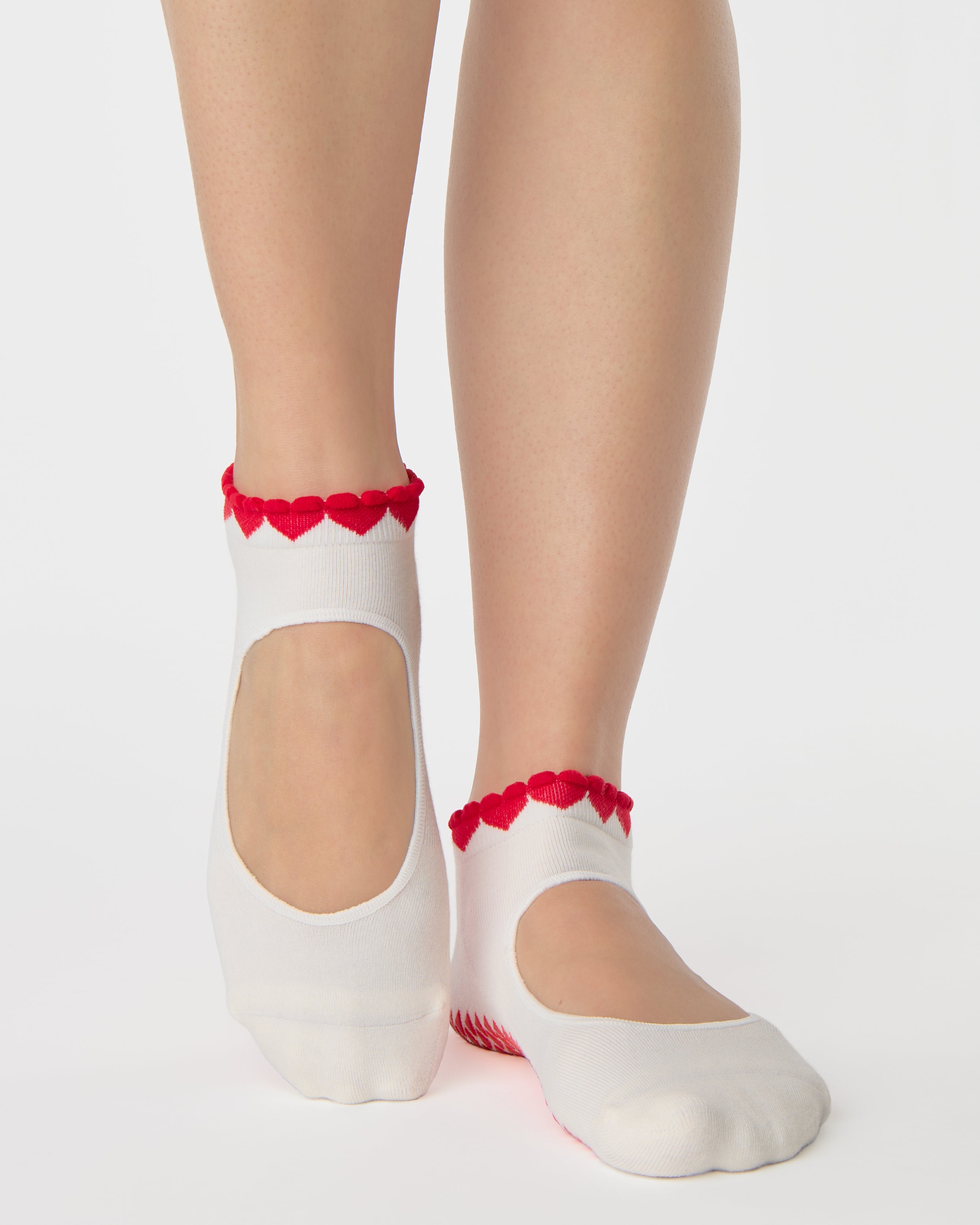 Woman wearing solid white strap grip sock with red heart wrapping around the scalloped cuff, made for pilates, barre, yoga, and beyond. 