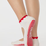 Woman wearing solid white strap grip sock with red heart wrapping around the scalloped cuff, made for pilates, barre, yoga, and beyond. 