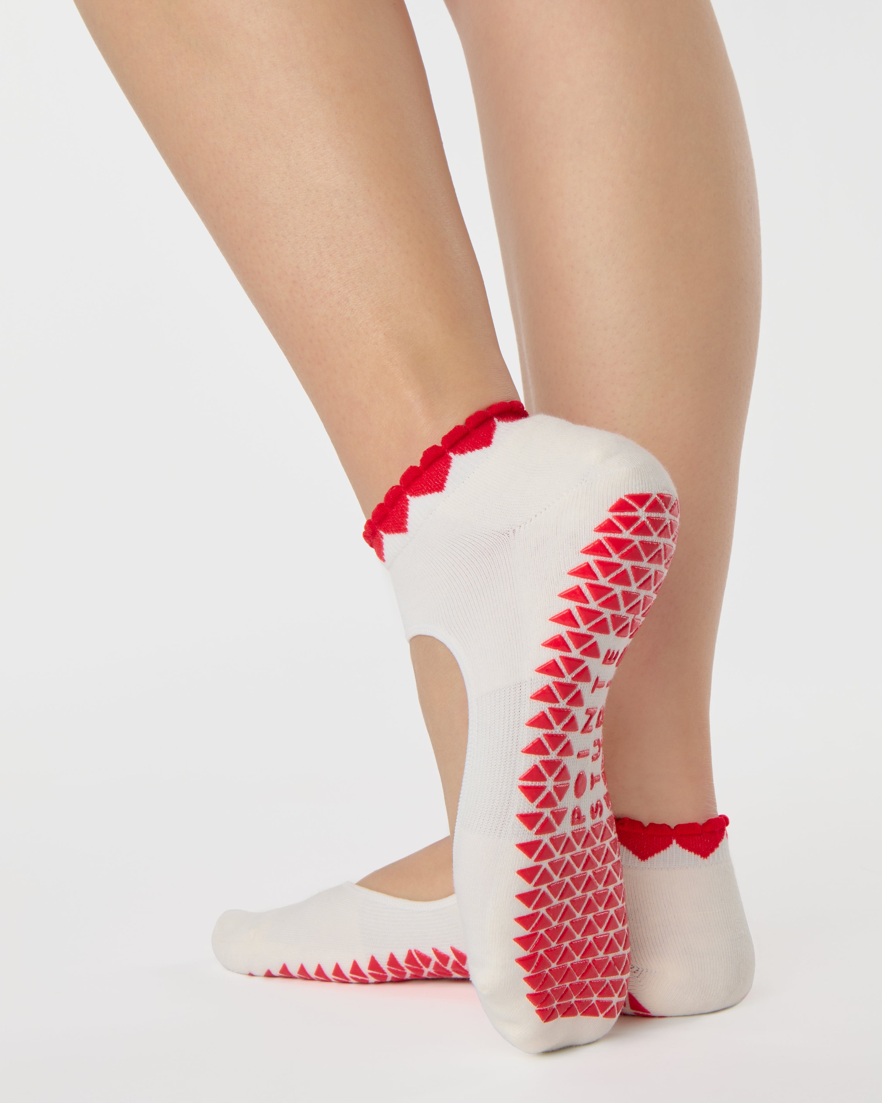 Woman wearing solid white strap grip sock with red heart wrapping around the scalloped cuff, made for pilates, barre, yoga, and beyond. 