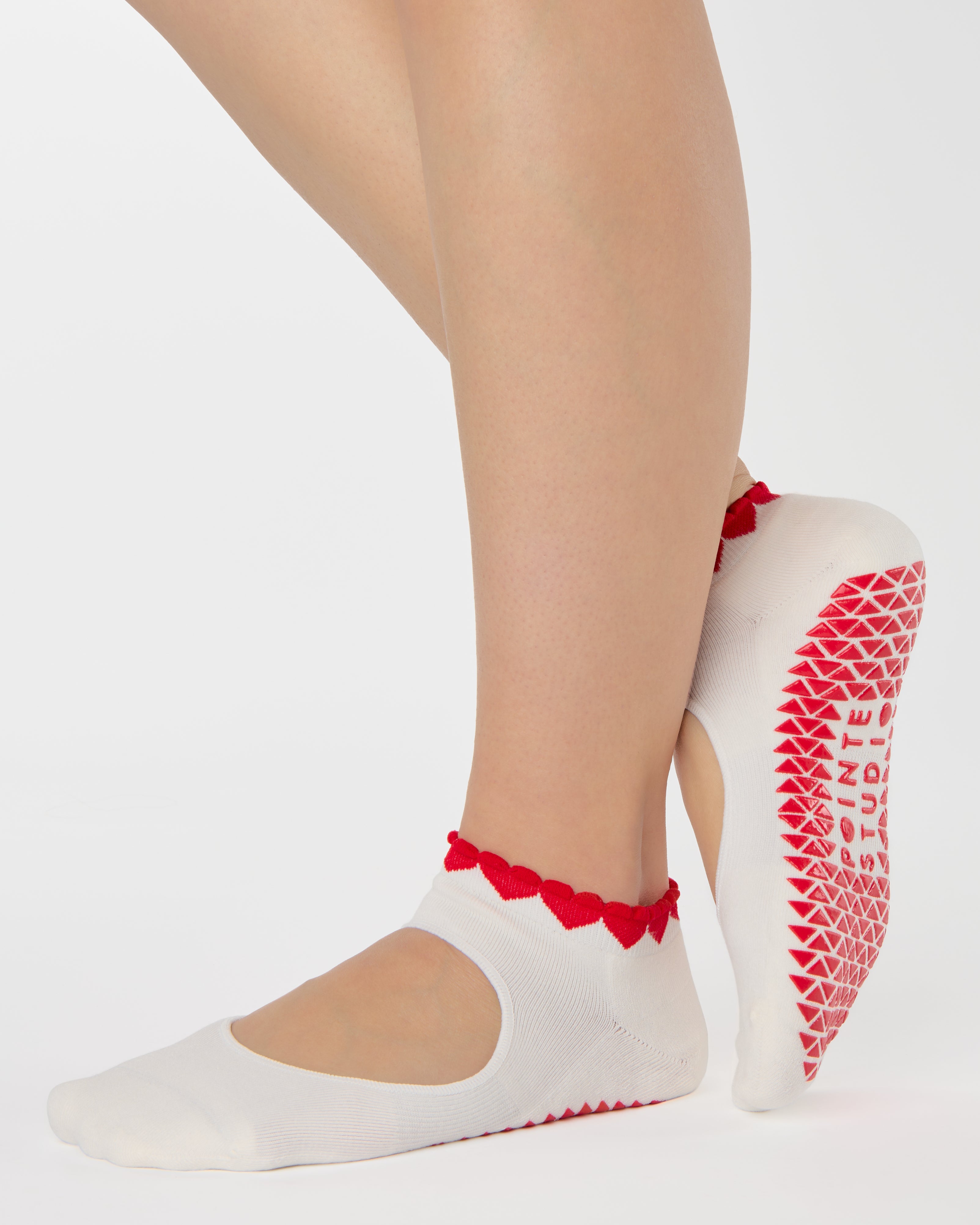 Woman wearing solid white strap grip sock with red heart wrapping around the scalloped cuff, made for pilates, barre, yoga, and beyond. 