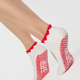 Woman wearing solid white strap grip sock with red heart wrapping around the scalloped cuff, made for pilates, barre, yoga, and beyond. 