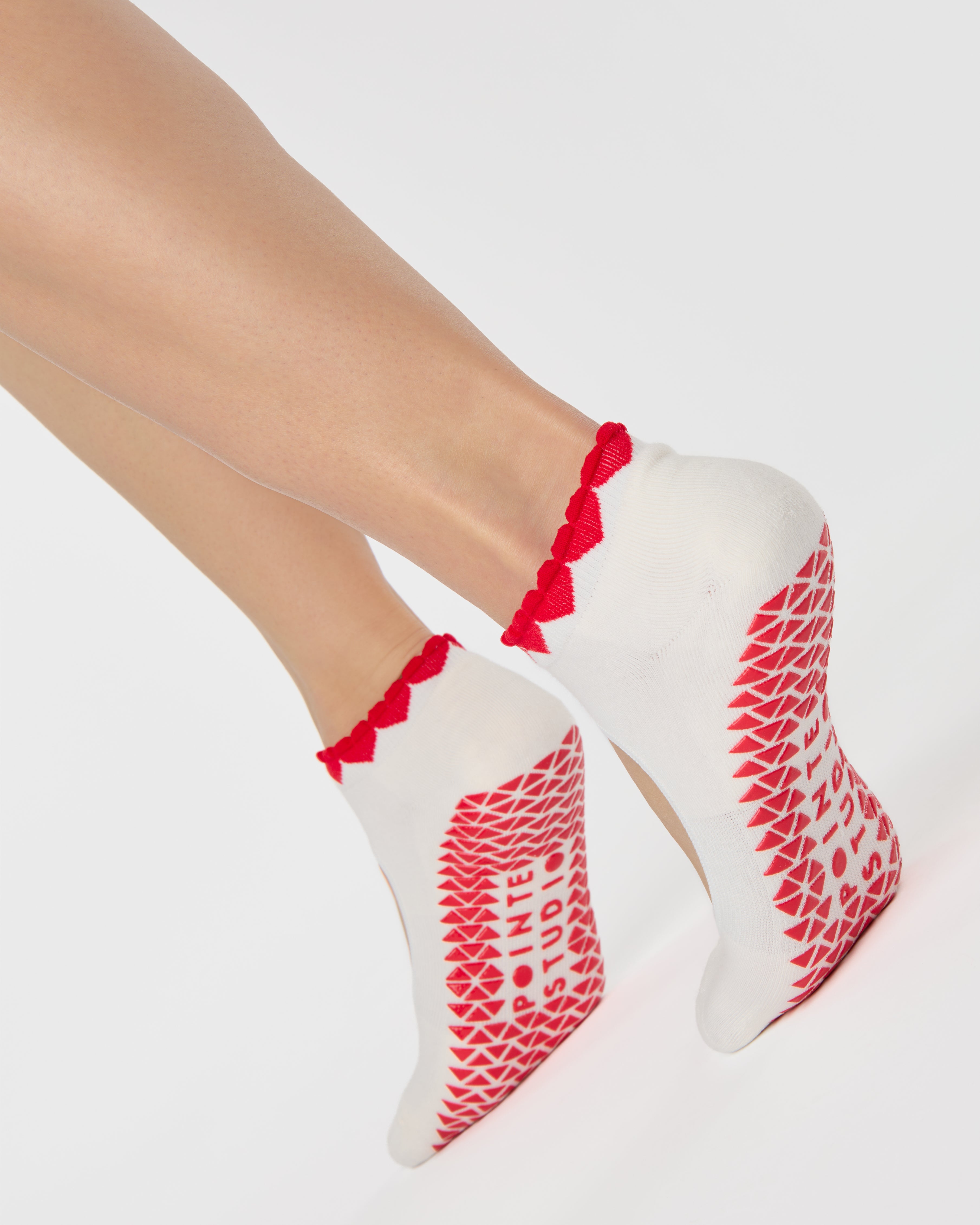 Woman wearing solid white strap grip sock with red heart wrapping around the scalloped cuff, made for pilates, barre, yoga, and beyond. 
