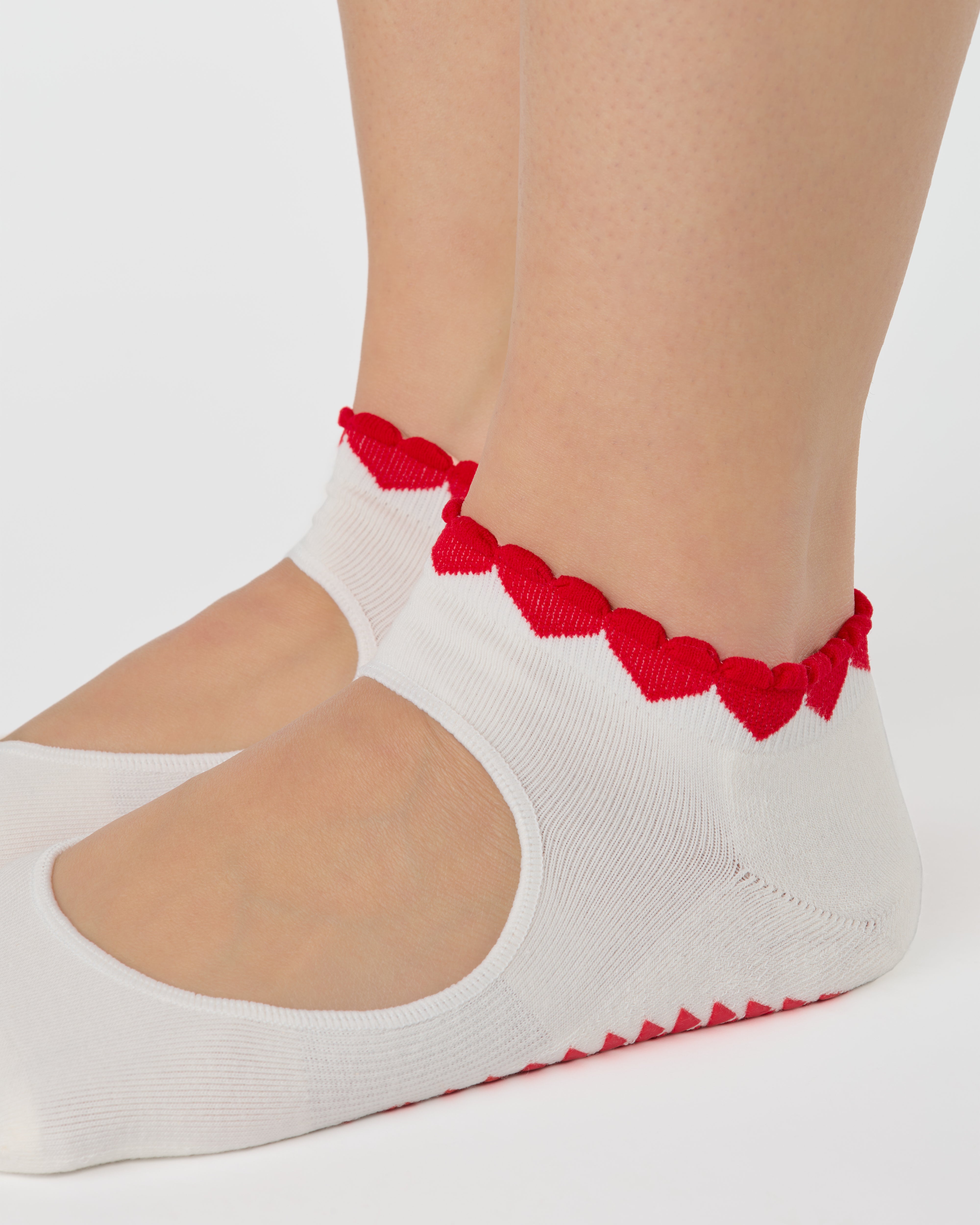 Woman wearing solid white strap grip sock with red heart wrapping around the scalloped cuff, made for pilates, barre, yoga, and beyond. 