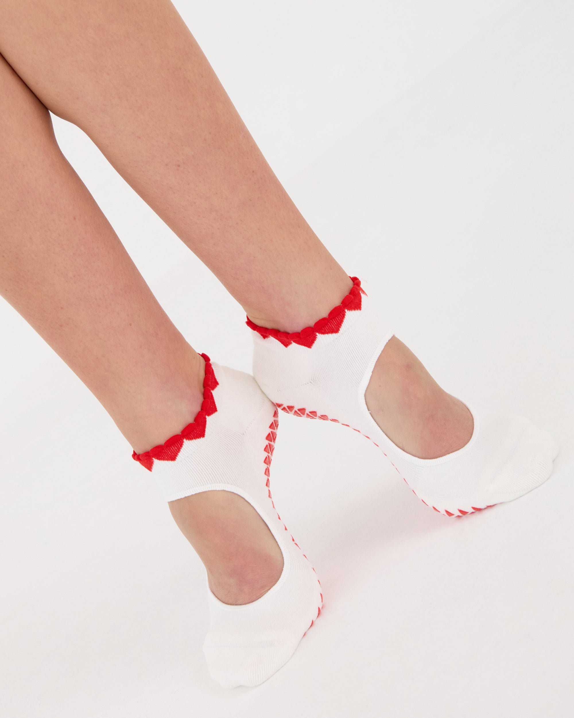 Woman wearing solid white strap grip sock with red heart wrapping around the scalloped cuff, made for pilates, barre, yoga, and beyond. 